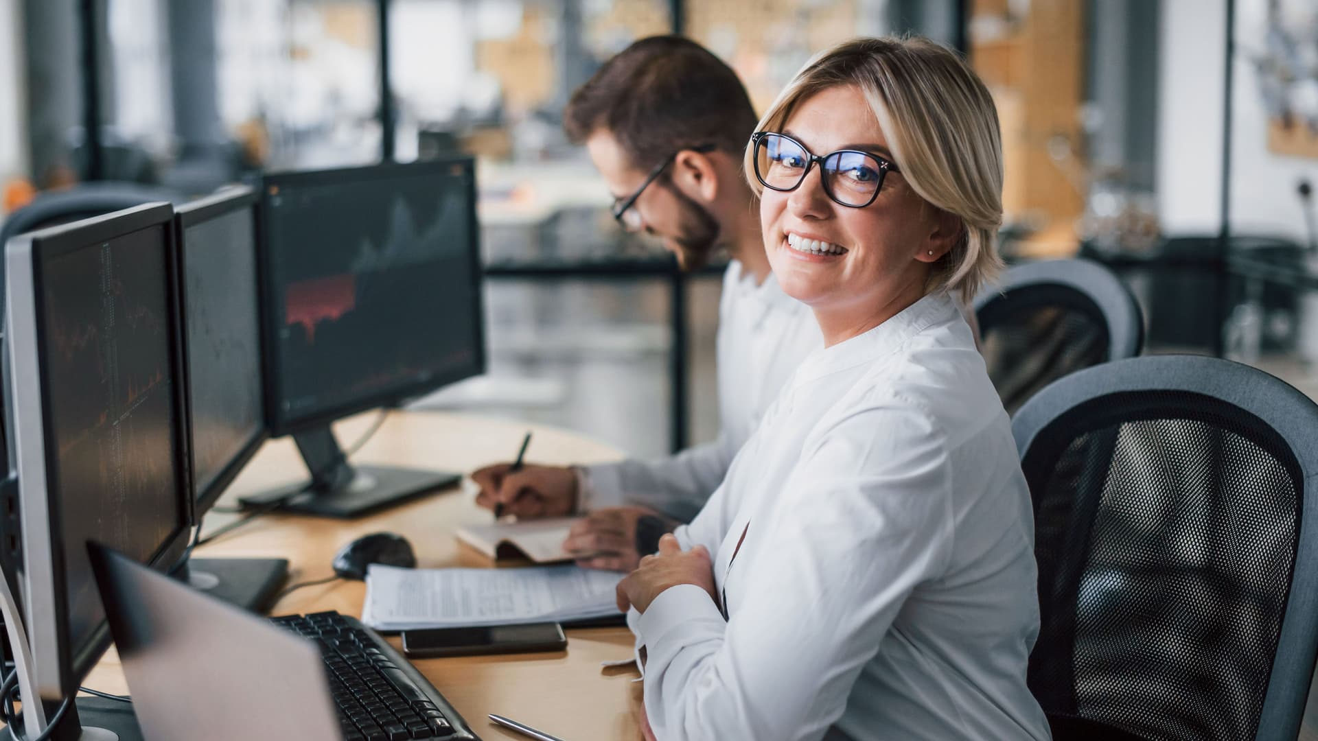 Eine Frau, die ihre Arme am Bürotisch aufstützt. Sie schaut in die Kamera. Im Hintergrund ist ein Kollege, der sich Notizen macht. Beide sitzen vor Computerbildschirmen.
