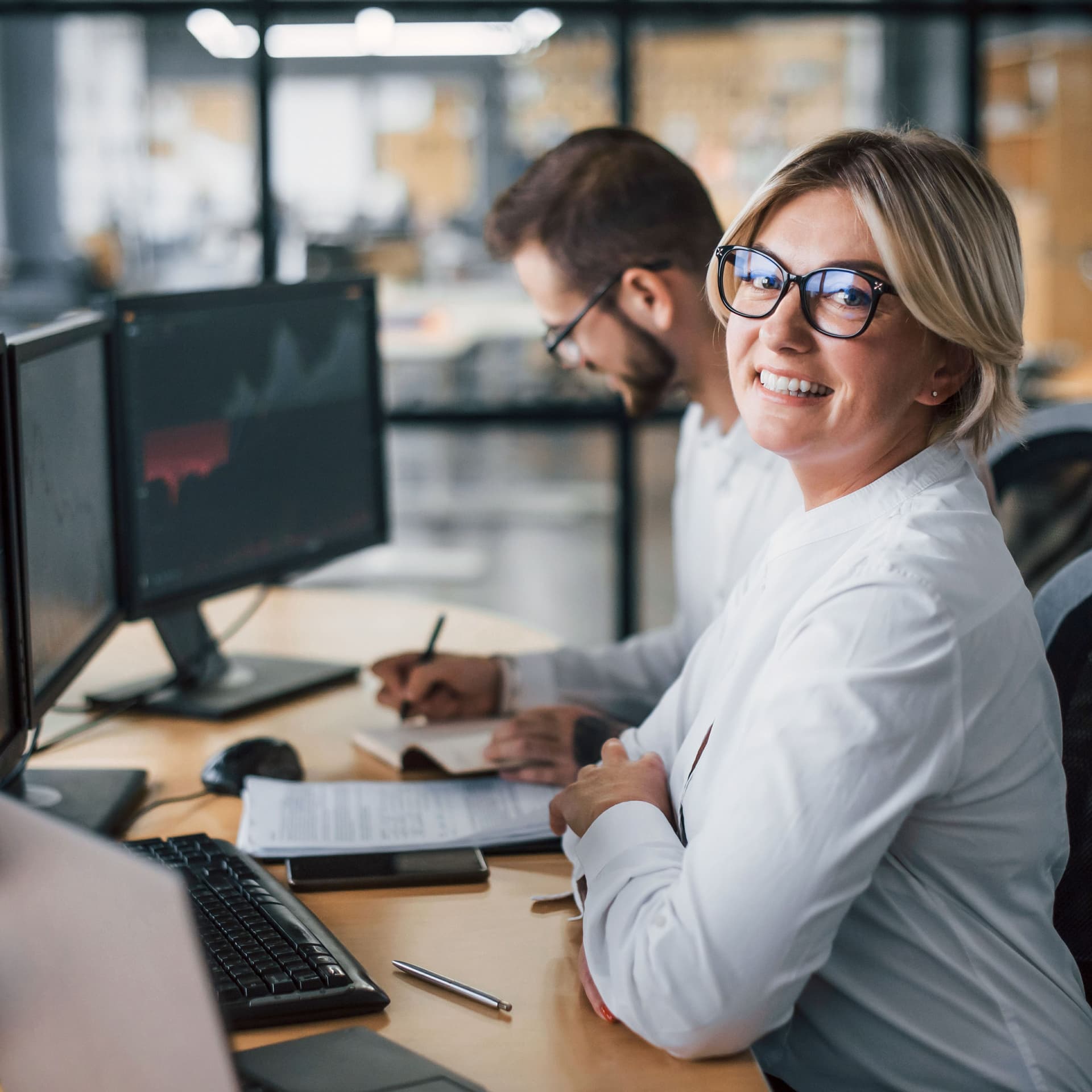 Eine Frau, die ihre Arme am Bürotisch aufstützt. Sie schaut in die Kamera. Im Hintergrund ist ein Kollege, der sich Notizen macht. Beide sitzen vor Computerbildschirmen.