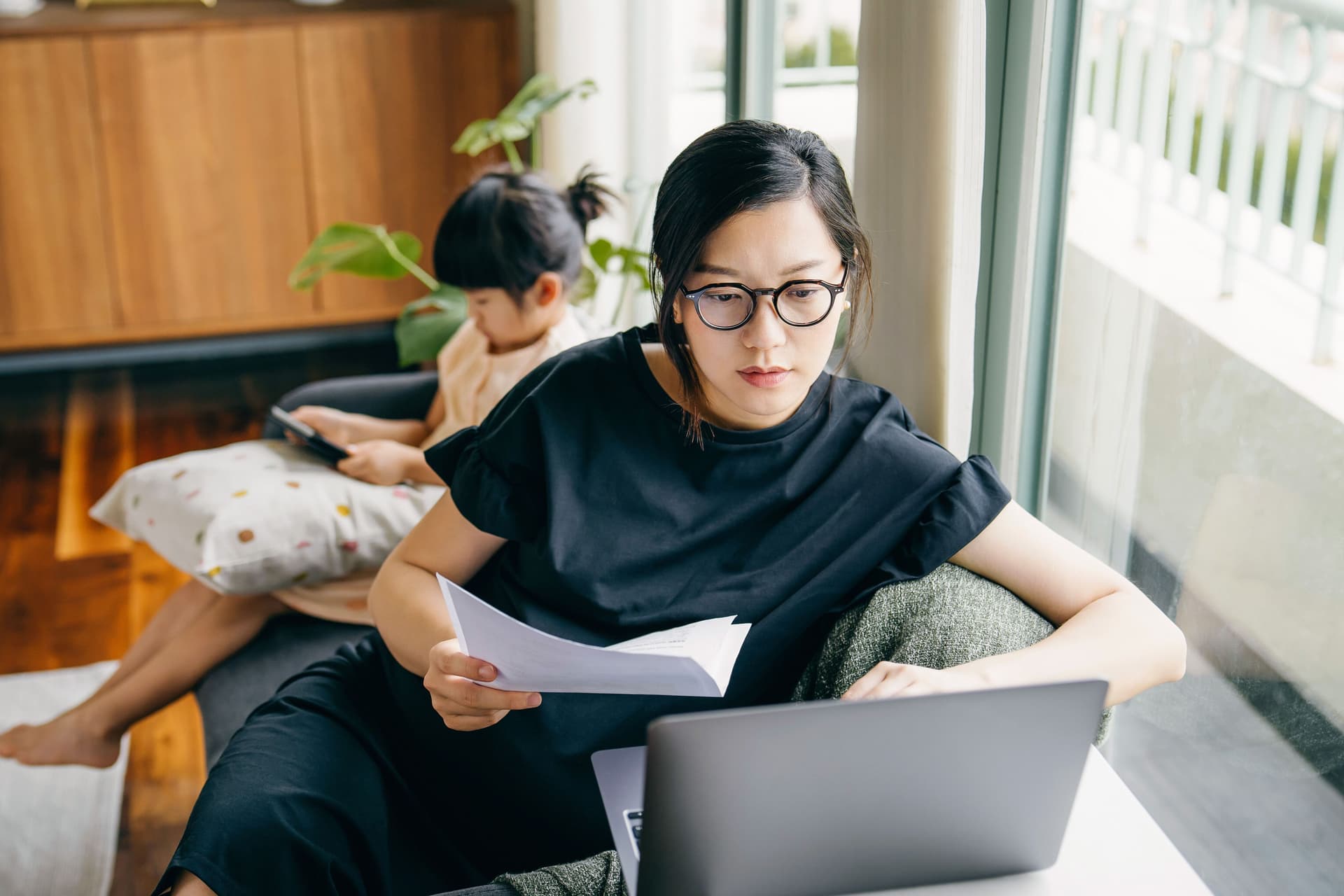 Junge Frau mit Brille und Papier in der Hand sitzt an einem Laptop. Im Hintergrund sitzt ein Kind mit einem Tablet.
