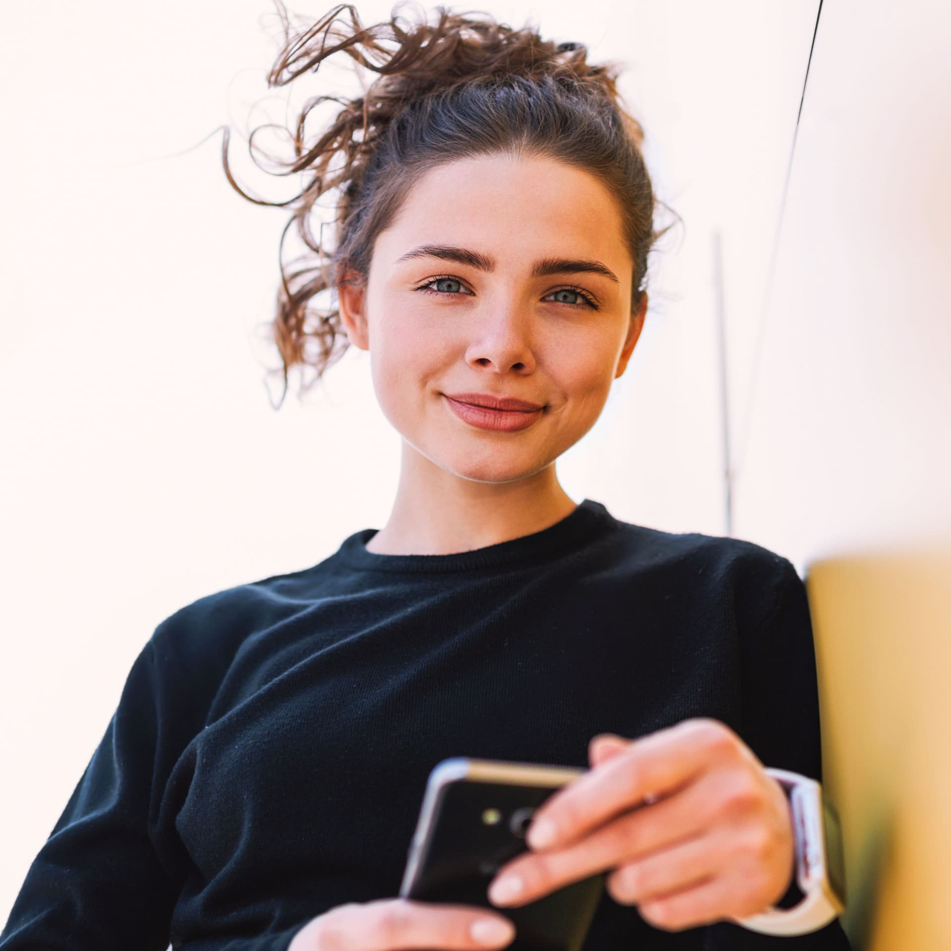Junge Frau lehnt mit Handy an einer Wand und schaut in die Kamera.