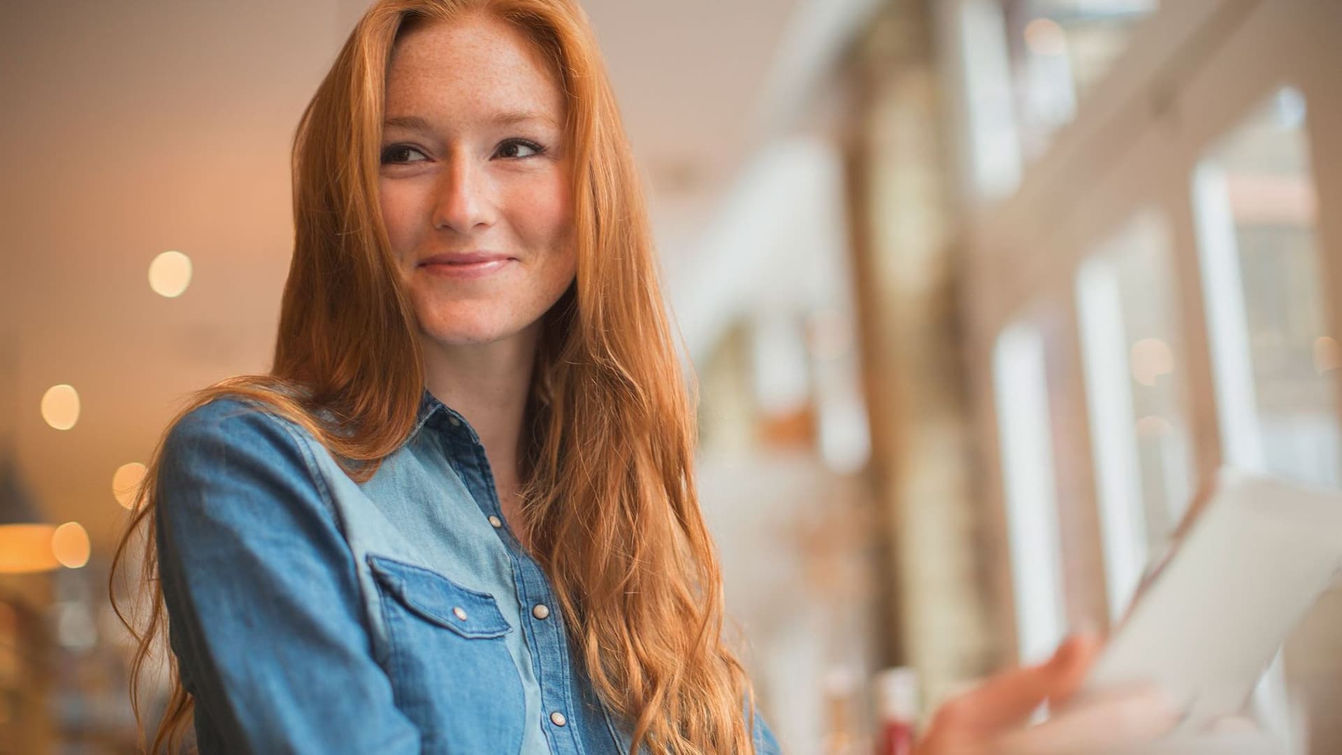 Eine junge Frau sitzt in einem Cafe und lächelt an der Kamera vorbei. Sie hat ein Tablet vor sich.