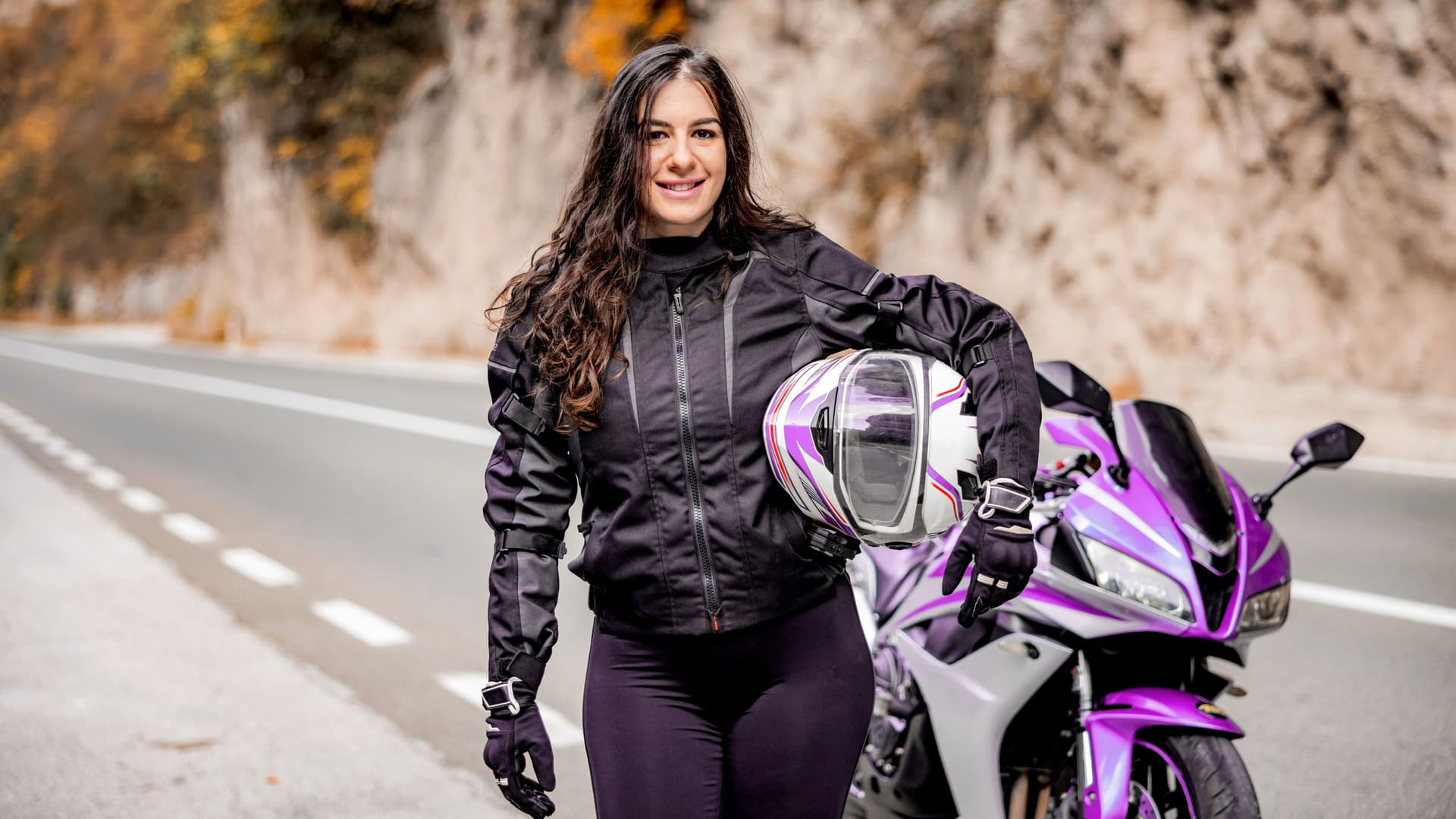 Junge Frau mit Helm unterm arm steht vor einem lilafarbenen Motorrad an einer Landstraße und lächelt in die Kamera.