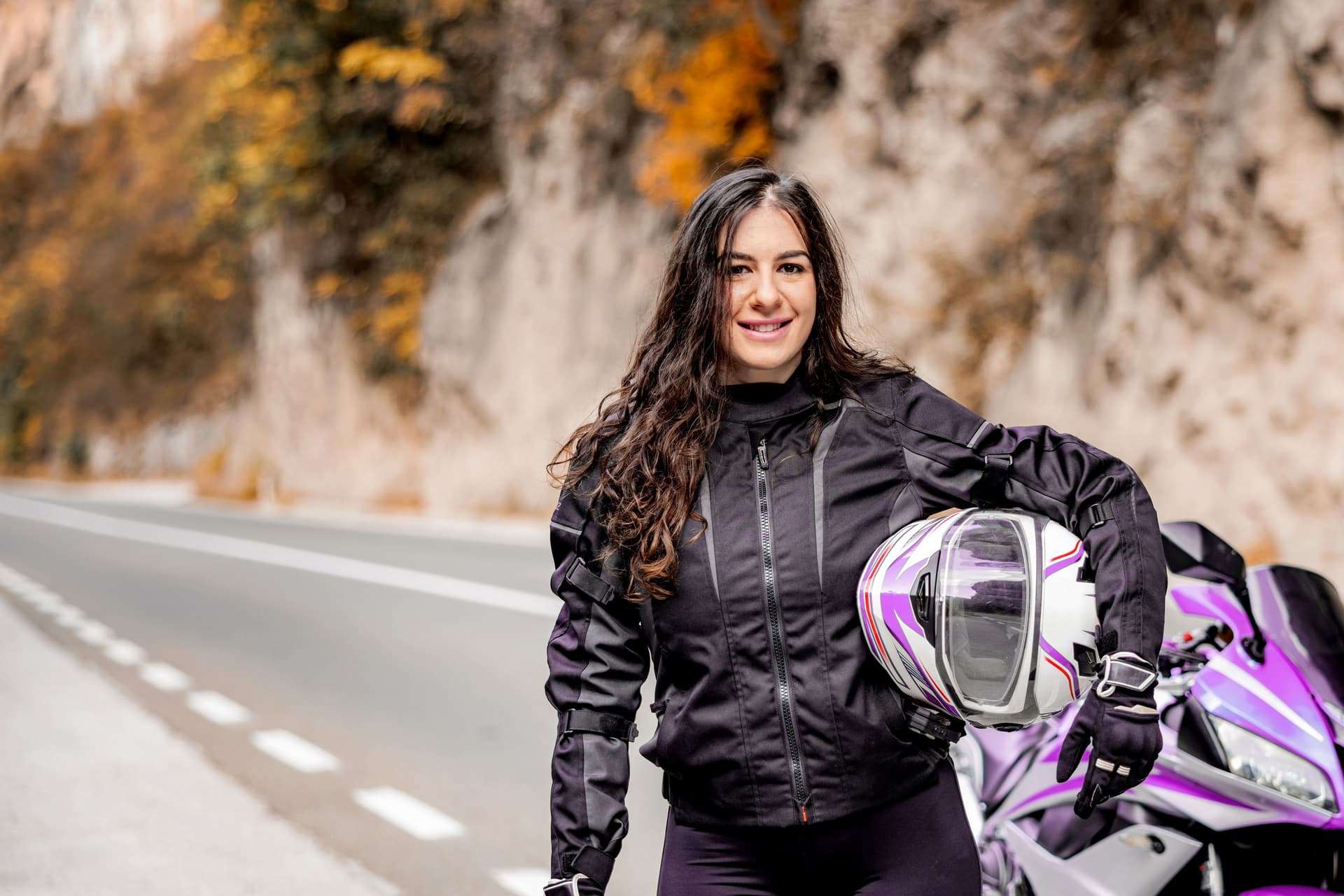Junge Frau mit Helm unterm arm steht vor einem lilafarbenen Motorrad an einer Landstraße und lächelt in die Kamera.