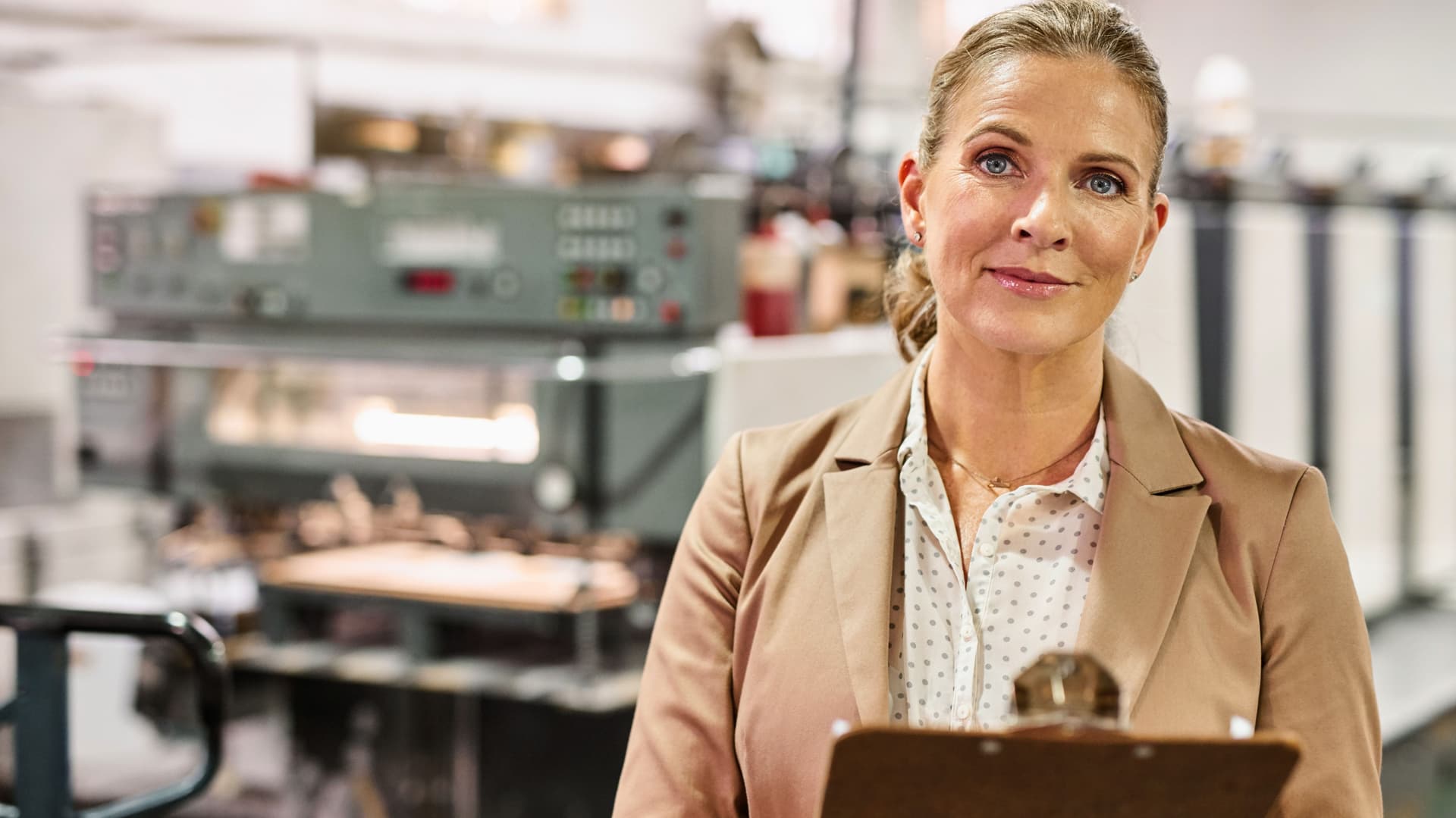 Frau im Trenchcoat mit Klemmbrett in einer Fabrik.