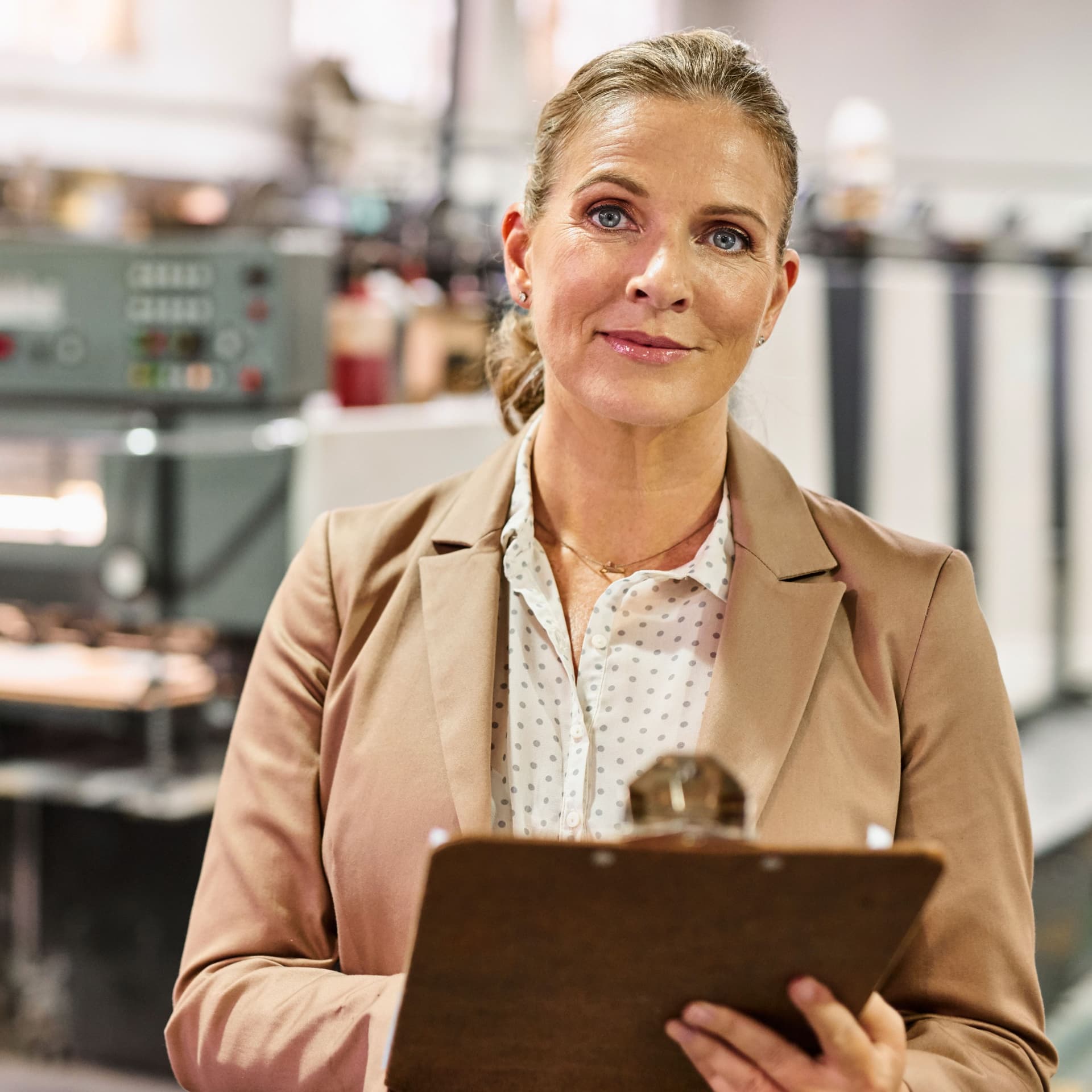 Frau im Trenchcoat mit Klemmbrett in einer Fabrik.