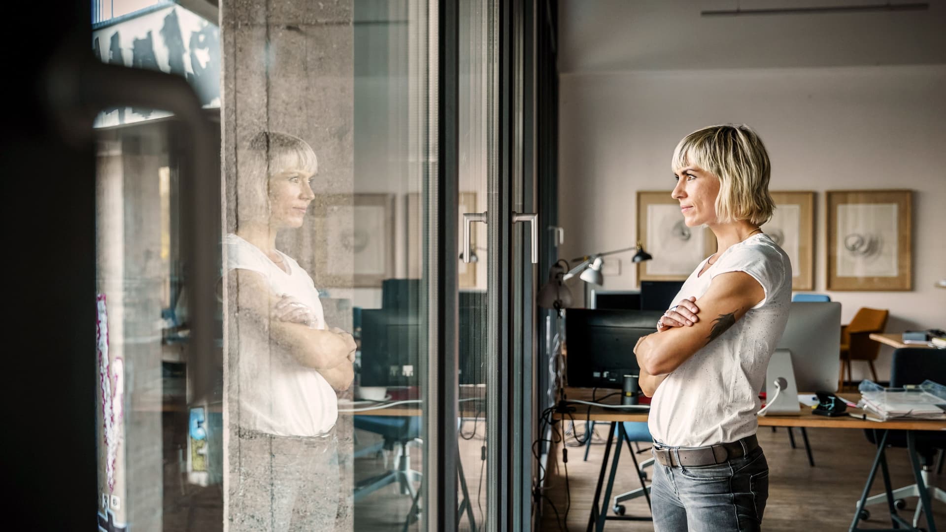 Junge Frau steht mit verschränkten Armen am Fenster eines Büros und schaut hinaus. Ihre Spiegelung ist im Glas zu erkennen.