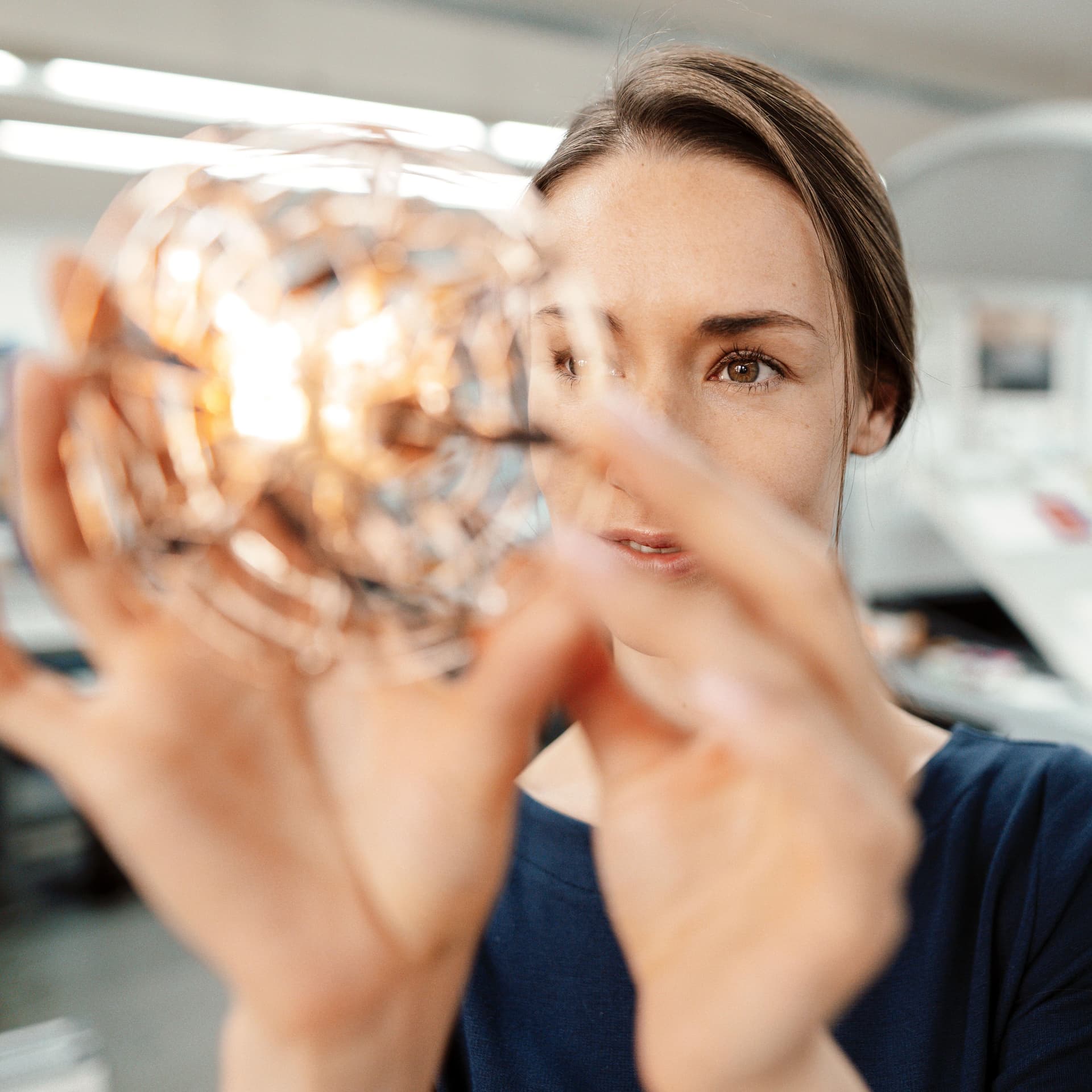 Junge Frau hält durchsichtigen, runden Gegenstand prüfend vors Gesicht in einem modernen Industrieumfeld.