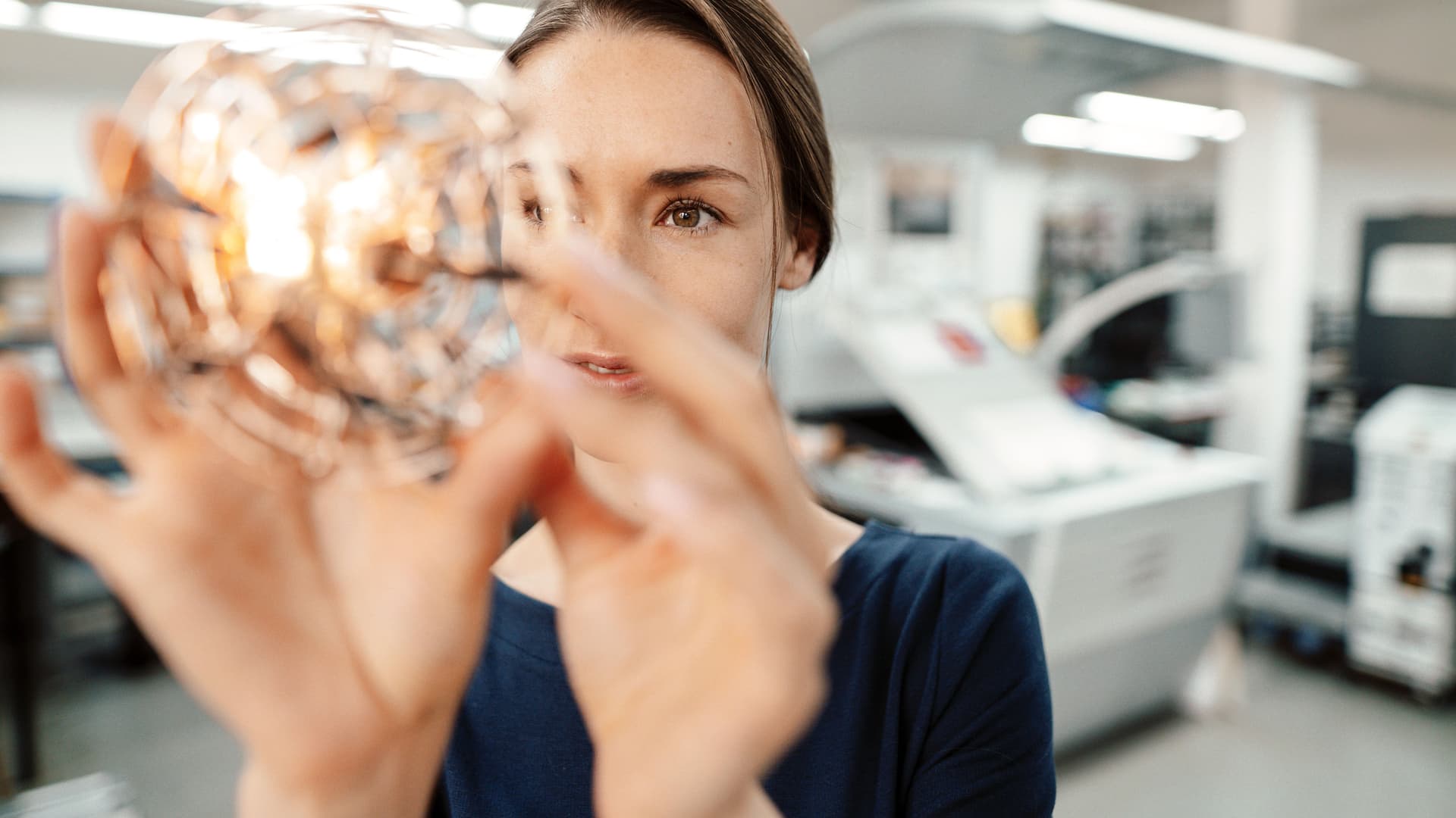 Junge Frau hält durchsichtigen, runden Gegenstand prüfend vors Gesicht in einem modernen Industrieumfeld.