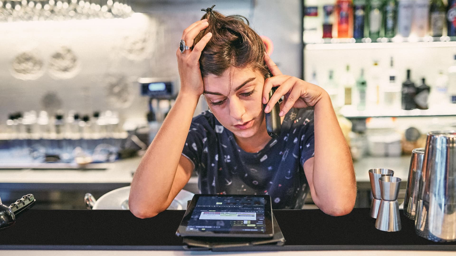 Frau hinter einer Theke schaut verzweifelt auf ein Tablet und telefoniert. Sie hat den Kopf auf eine Hand gestützt.