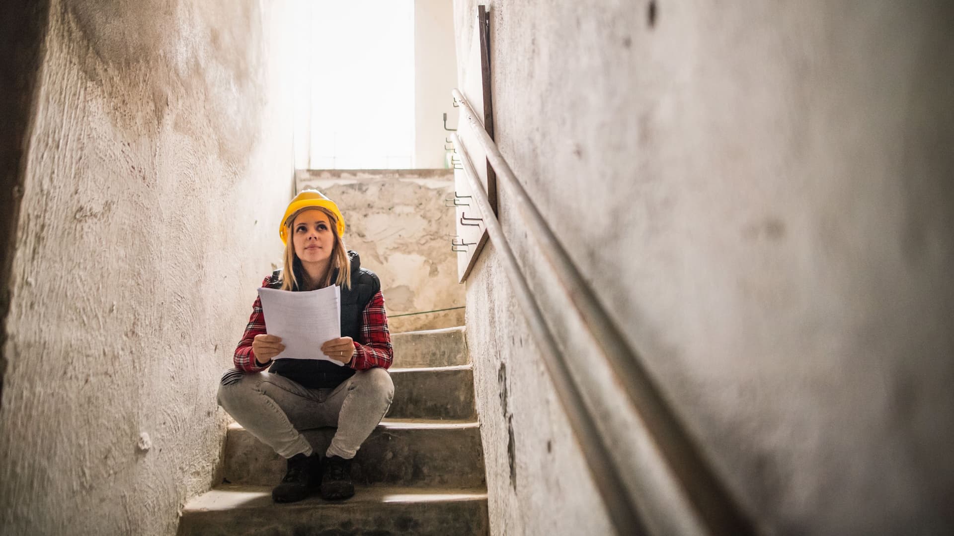 Junge Frau mit einem gelben Helm sitzt auf einer Steintreppe auf einer Baustelle. Sie hält Pläne in den Händen.