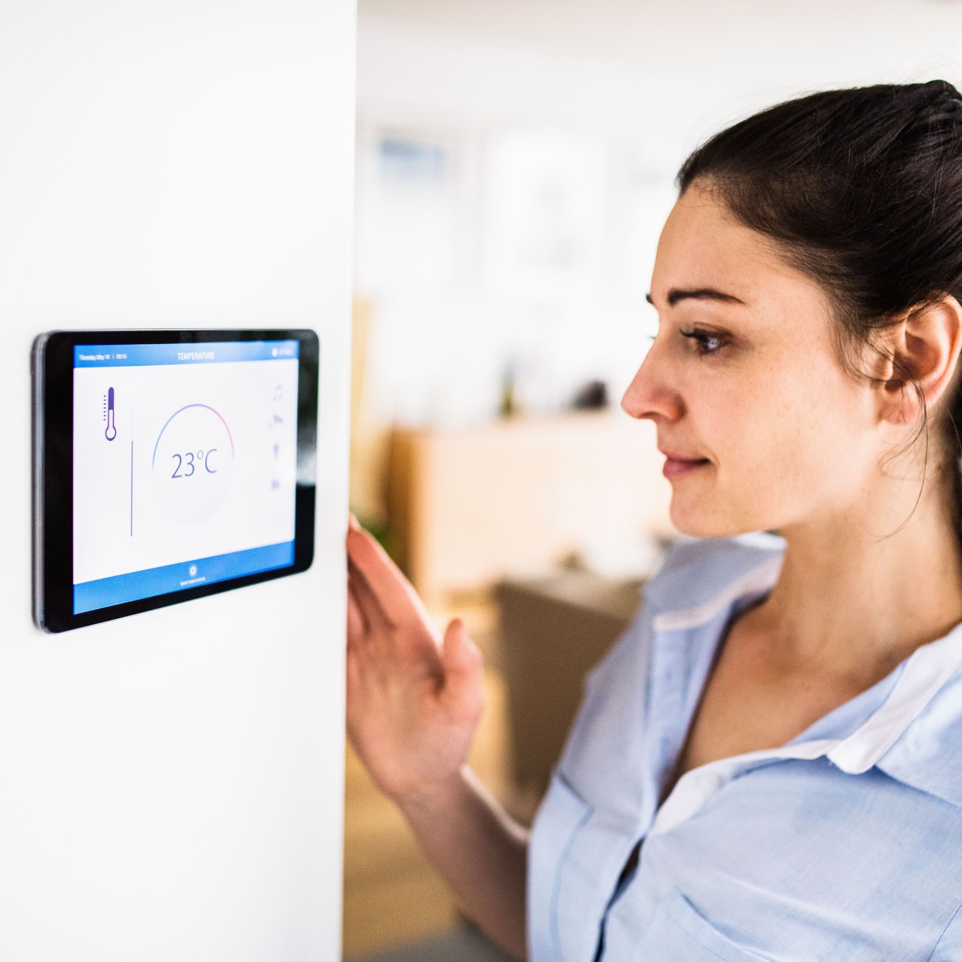 Eine Frau schaut auf ein Tablet an der Wand mit Smart Home-Bildschirm. Das Display zeigt die Temperatur 23°C.