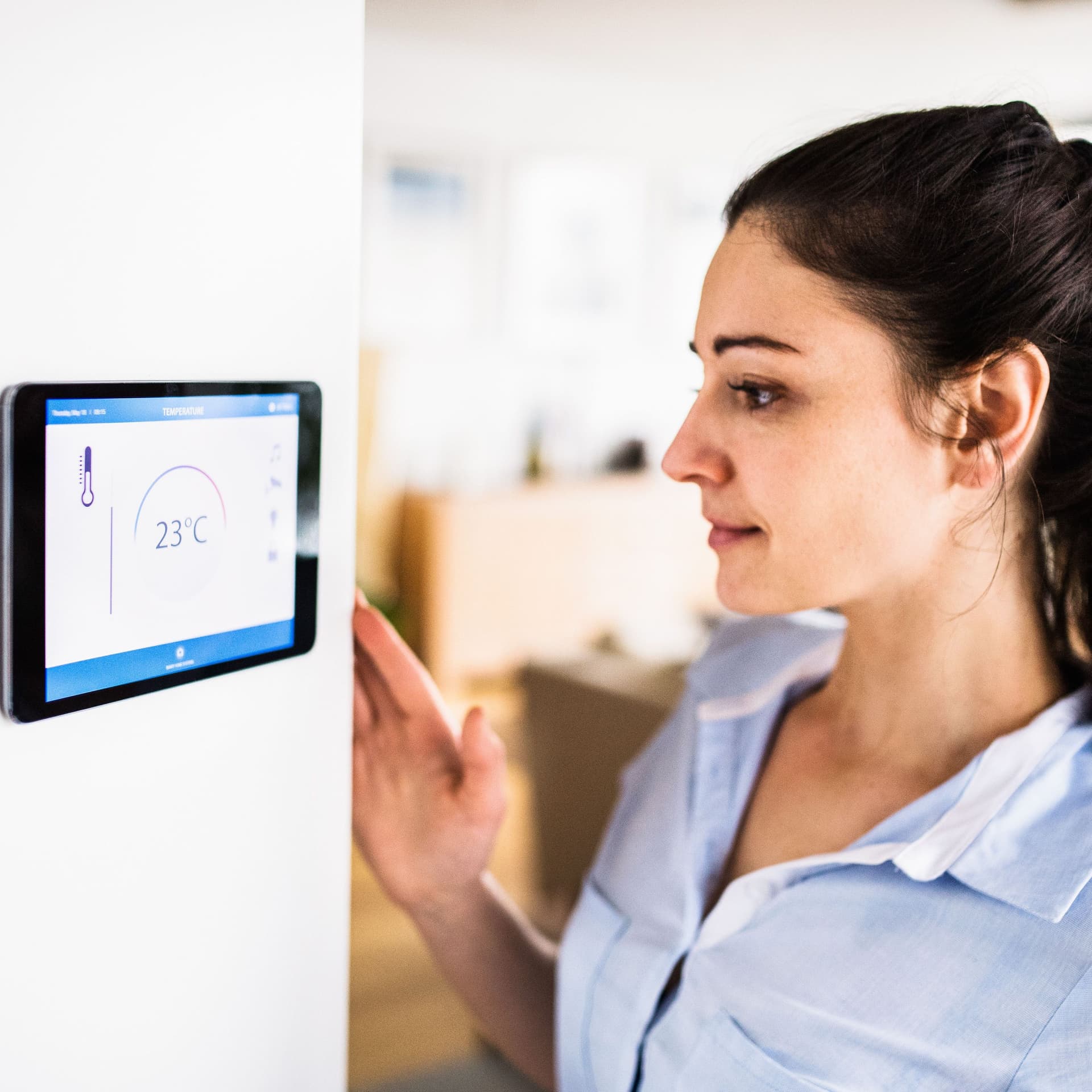 Eine Frau schaut auf ein Tablet an der Wand mit Smart Home-Bildschirm. Das Display zeigt die Temperatur 23°C.