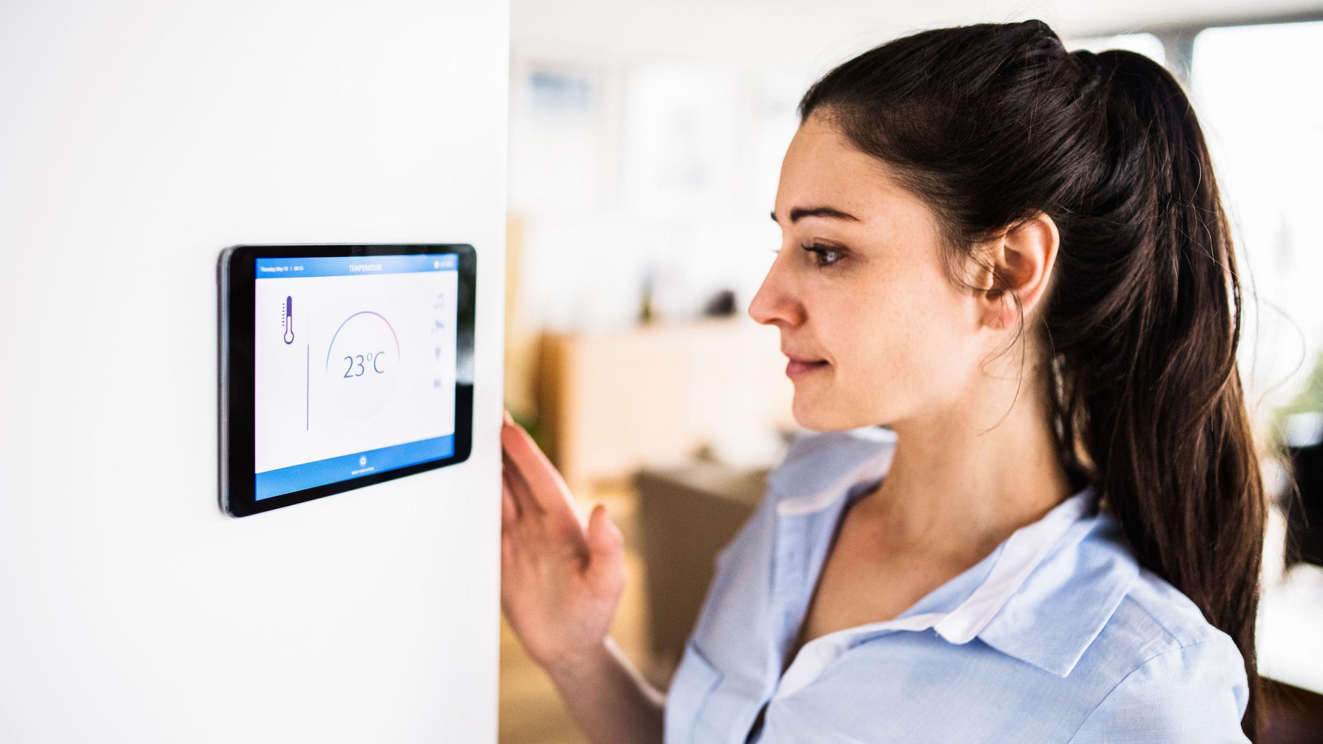 Eine Frau schaut auf ein Tablet an der Wand mit Smart Home-Bildschirm. Das Display zeigt die Temperatur 23°C.