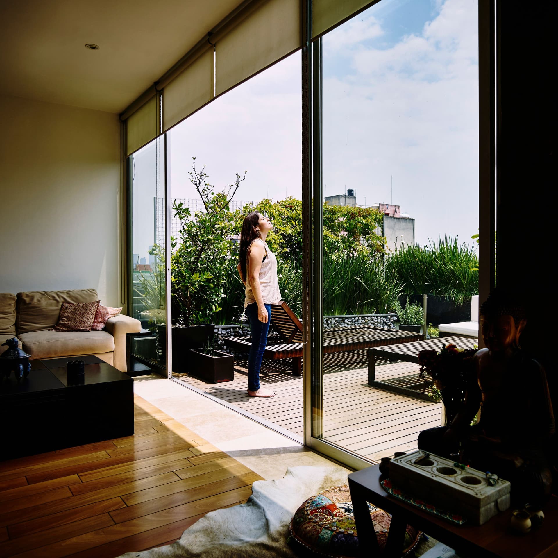 Blick aus einem Raum auf eine Frau, die ihr Gesicht zur Sonne hebt und auf einer großen Gartenterrasse steht. Die großen Panoramafenster sind geöffnet.