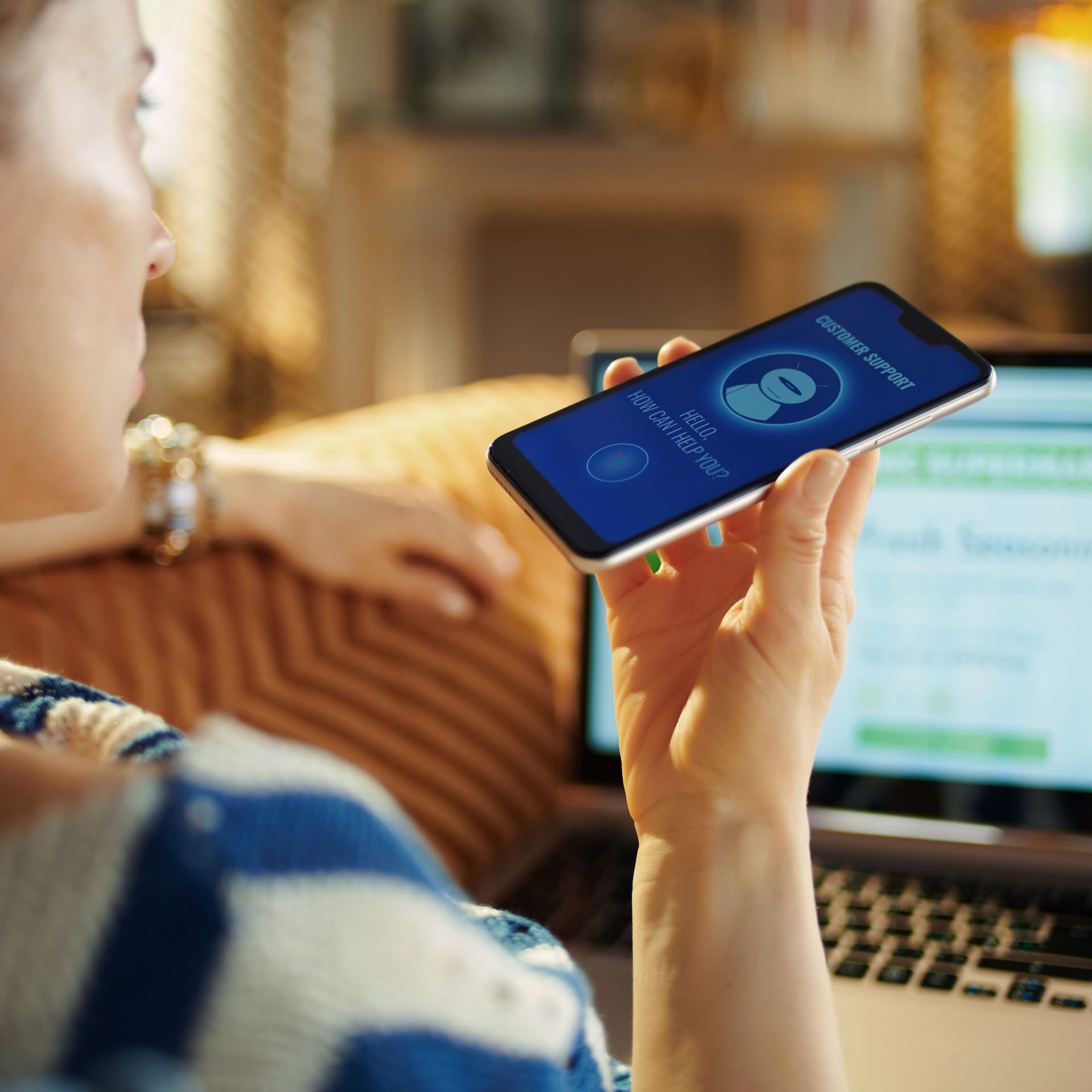 Von hinten über die Schulter fotografiert. Eine junge Frau auf der Couch verwendet den Sprachroboter ihres Smartphones.