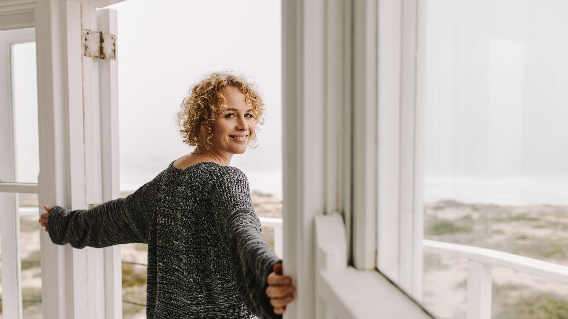 Seitenansicht einer Frau, die mit ausgebreiteten Armen in einer Balkontür steht und in die Kamera schaut. Vor ihr sind Dünen und das Meer.