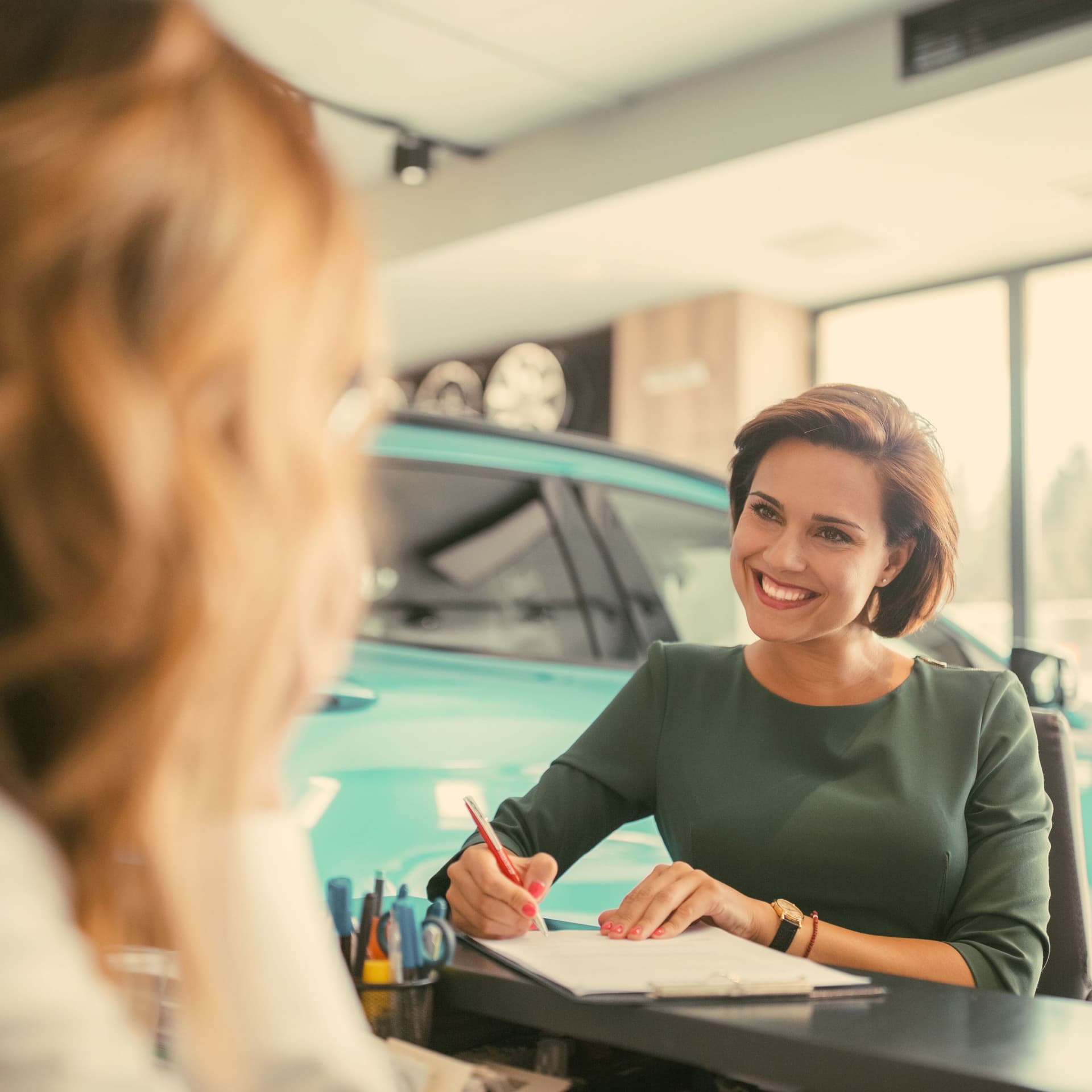 Zwei Frauen in einem Autohaus. Eine Frau unterschreibt lächelnd ein Dokument. 
