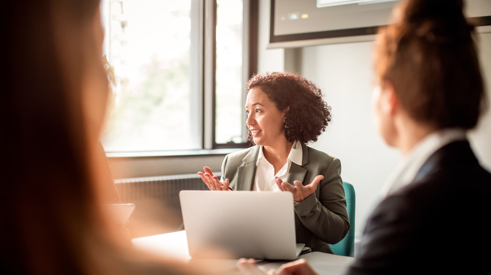 Mehrere Frauen sitzen zusammen bei einem Business Meeting in einem Konferenzraum. Man sieht eine Frau, die vor einem Laptop sitzt und mit den Händen gestikuliert.