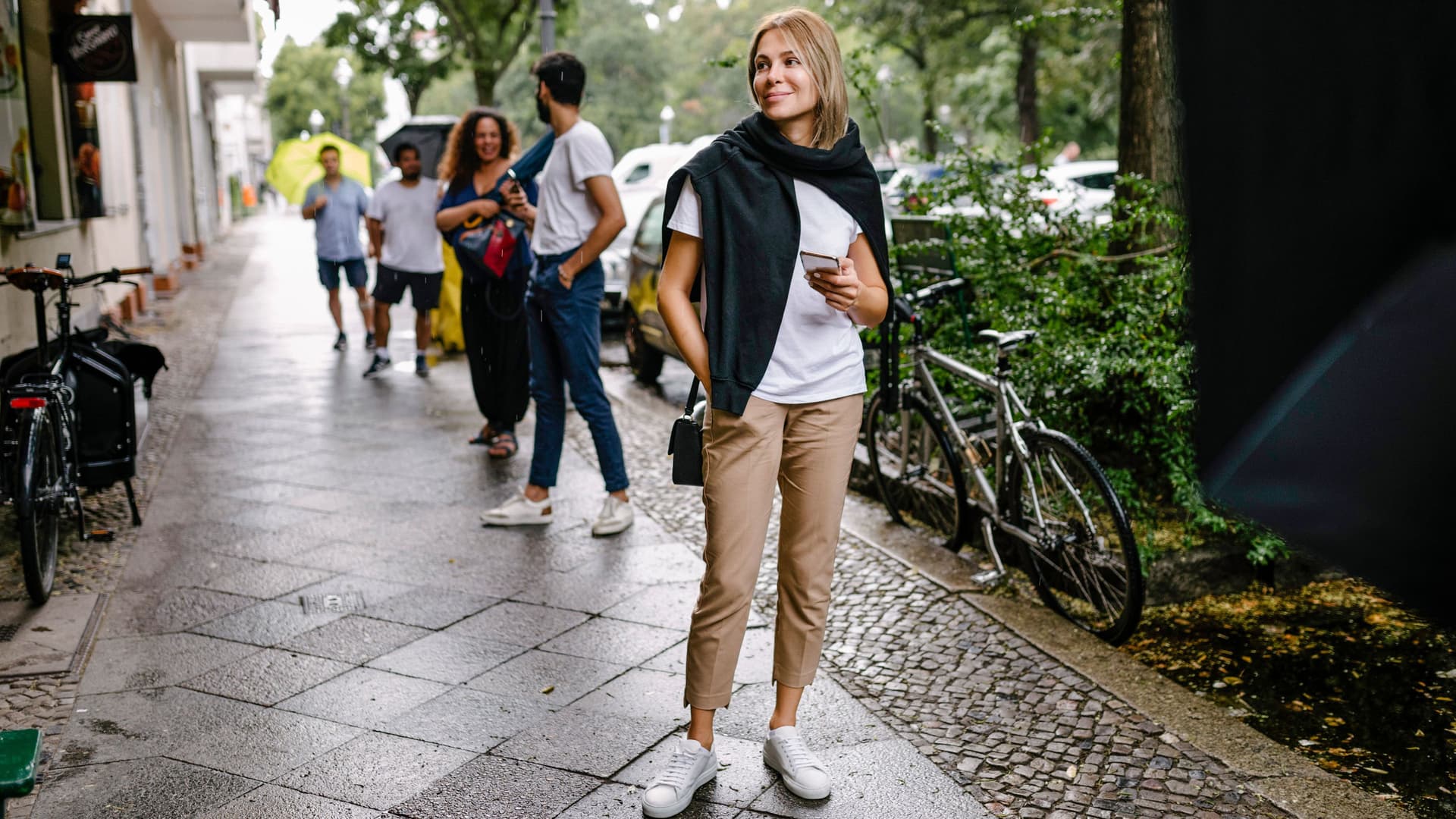 Menschen stehen mit Abstand auf dem Gehweg in einer Schlange an. Vorne steht eine junge Frau mit Smartphone in der Hand und lächelt. 