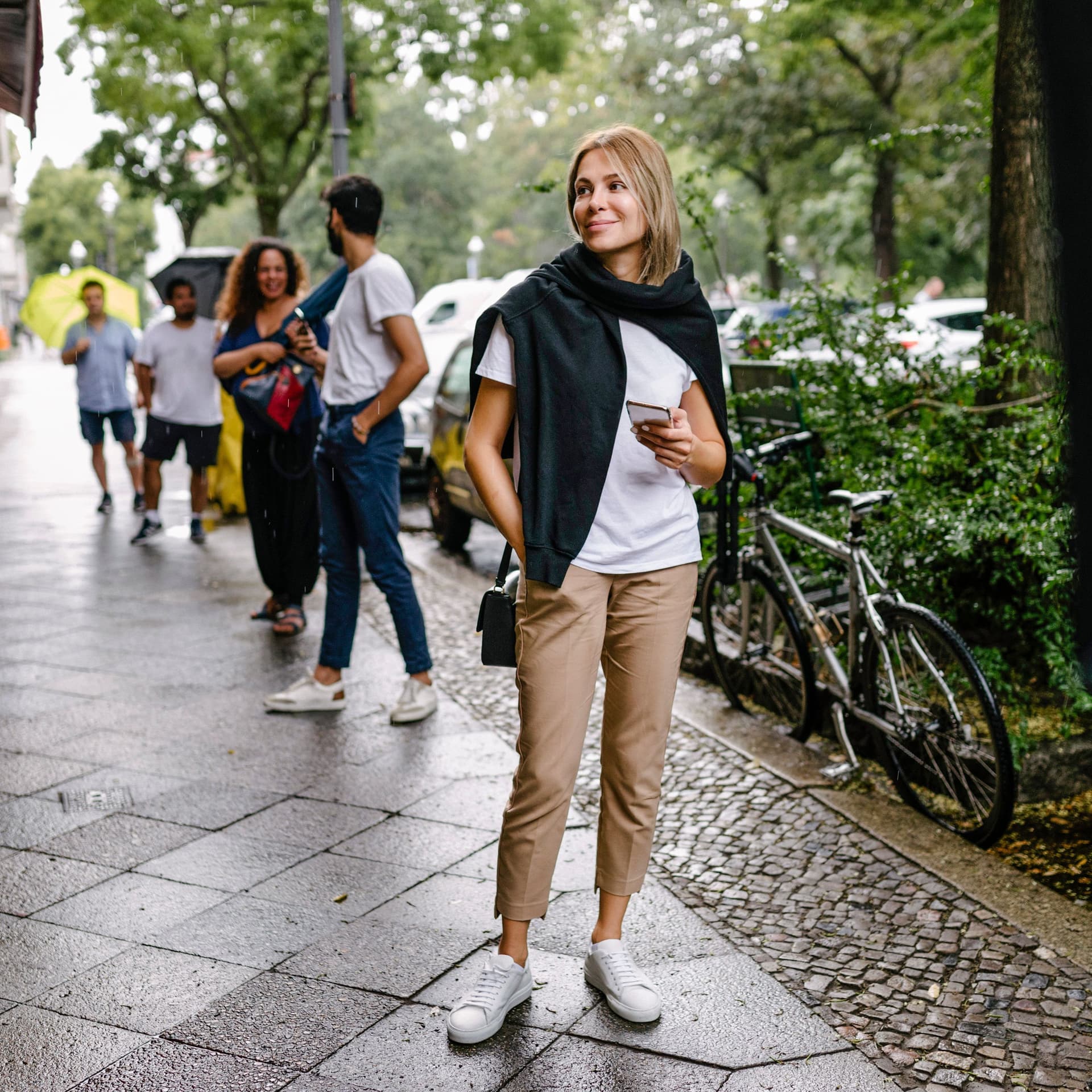Menschen stehen mit Abstand auf dem Gehweg in einer Schlange an. Vorne steht eine junge Frau mit Smartphone in der Hand und lächelt. 