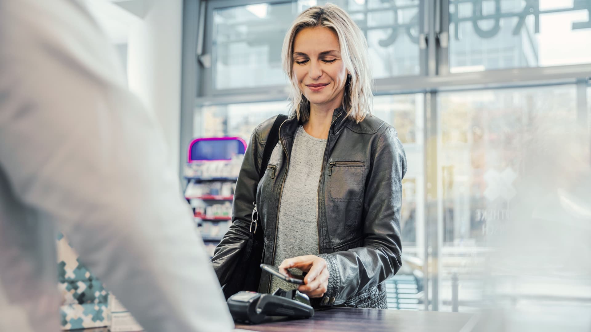 Lächelnde Kundin mittleren Alters beim Bezahlen mit dem Handy an der Kasse einer Apotheke