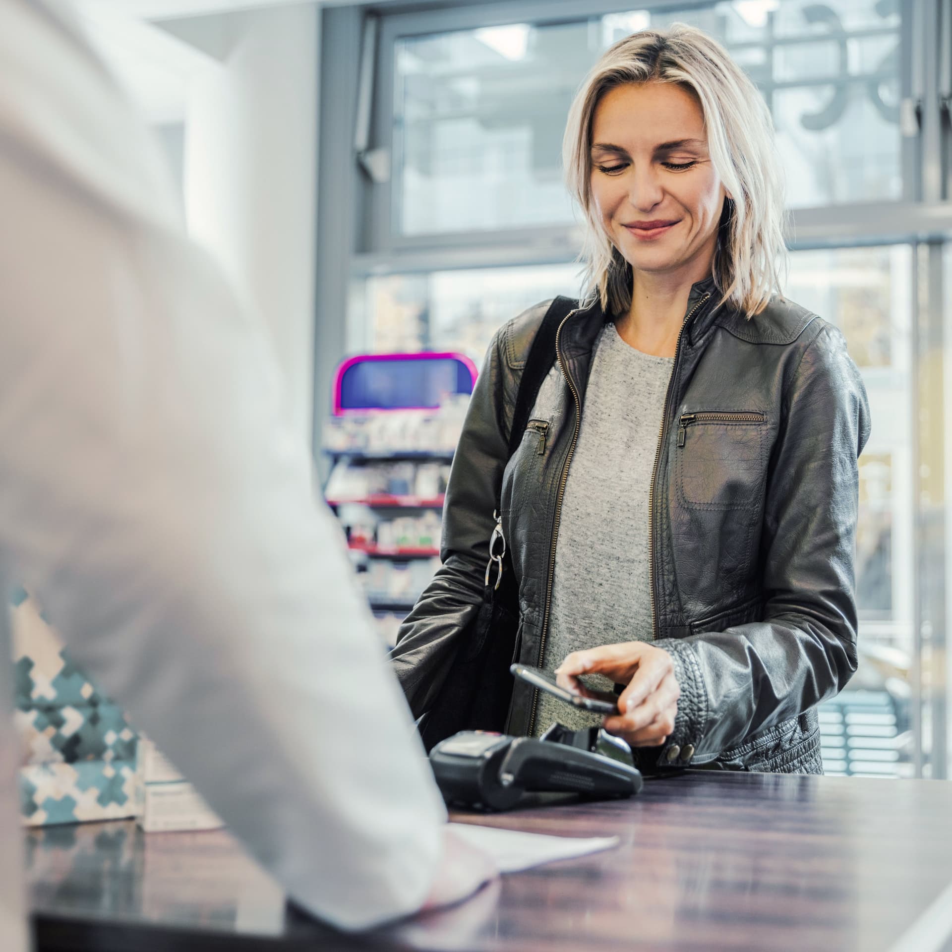 Lächelnde Kundin mittleren Alters beim Bezahlen mit dem Handy an der Kasse einer Apotheke
