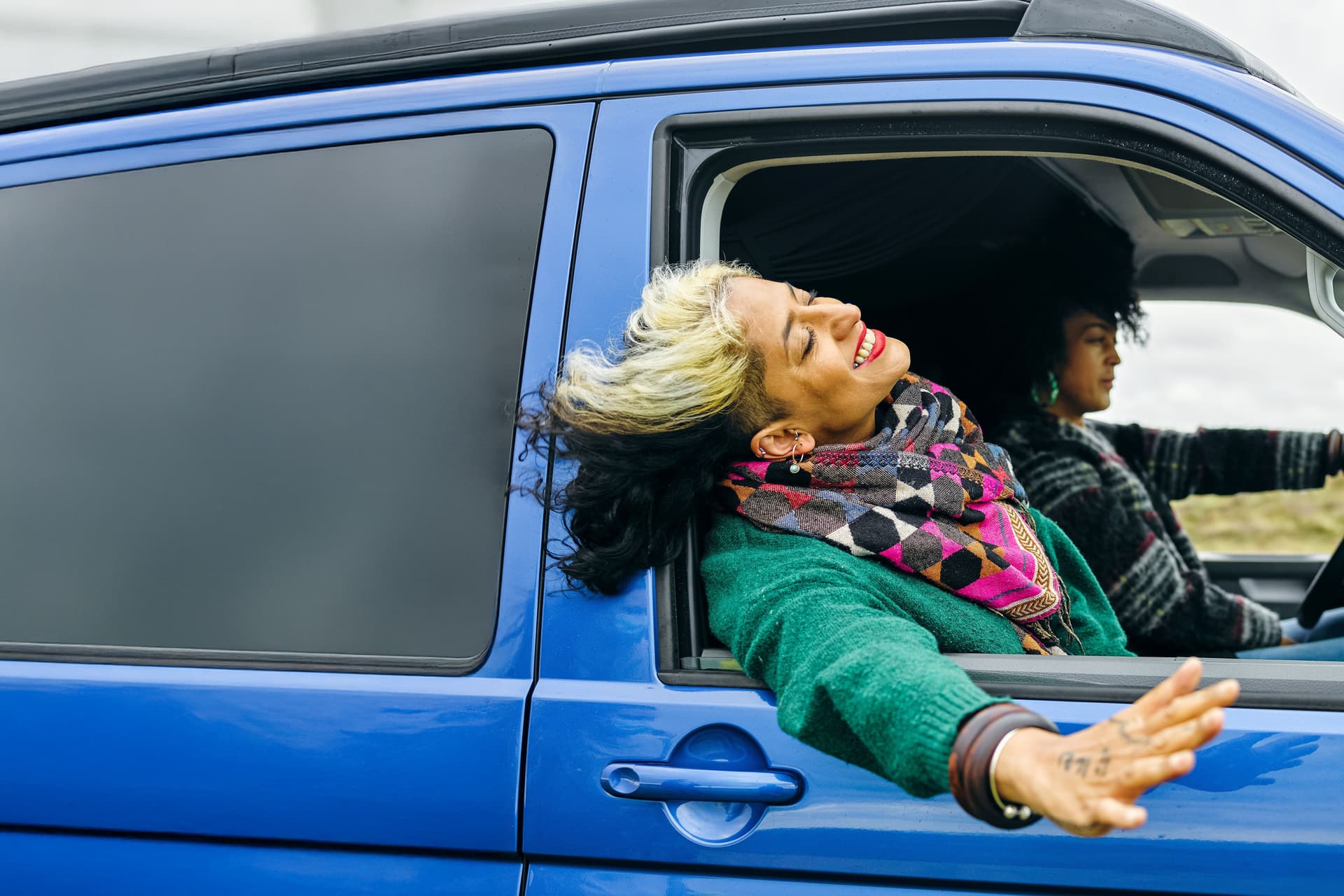 Zwei Frauen fahren in einem blauen Van. Das Fenster beim Beifahrersitz ist offen und die Frau streckt lachend ihren Kopf und ihre Hand aus dem Fenster.