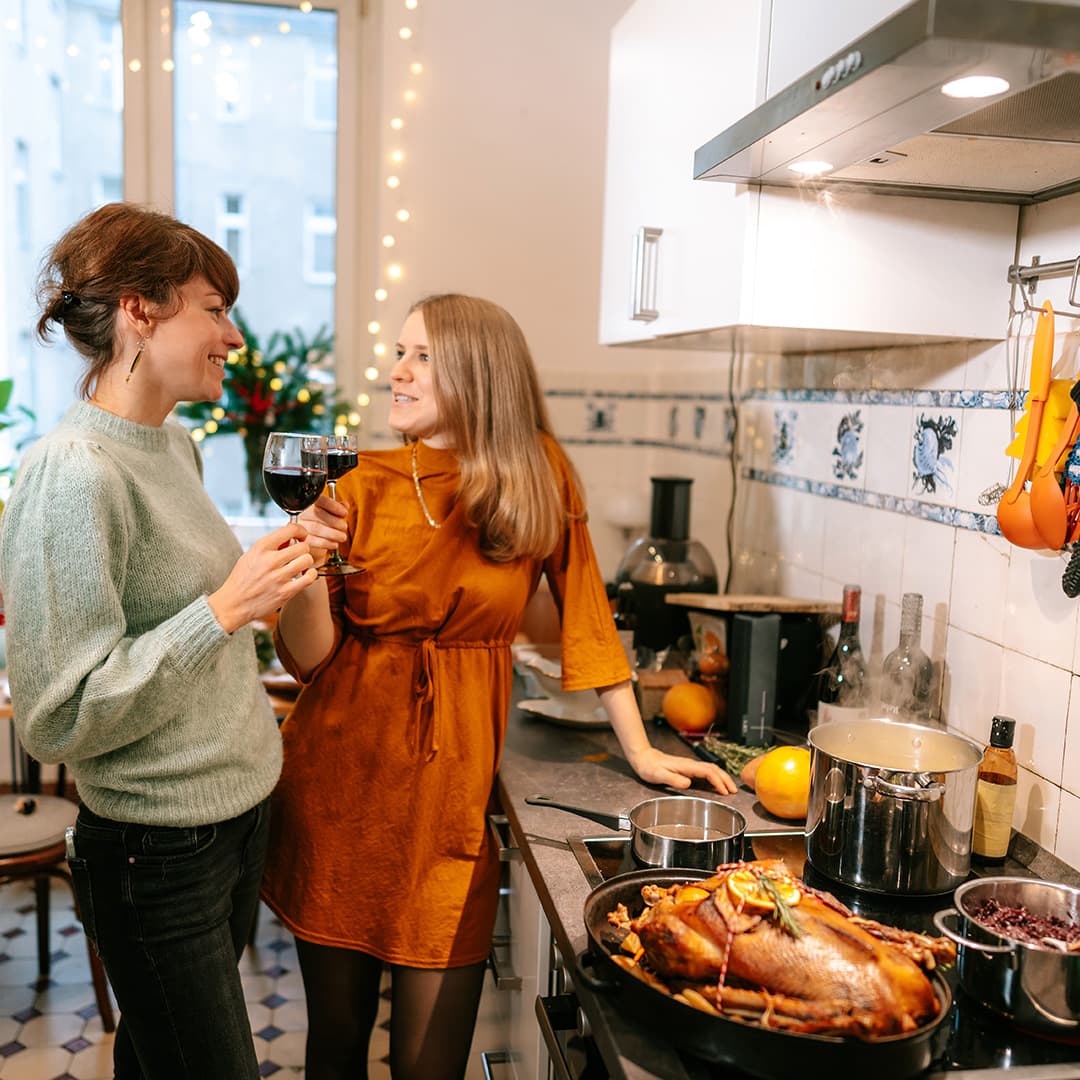 Frauen stehen mit einem Rotweinglas in der Küche. Sie unterhalten sich. Auf dem Herd steht eine Weihnachtsgans.