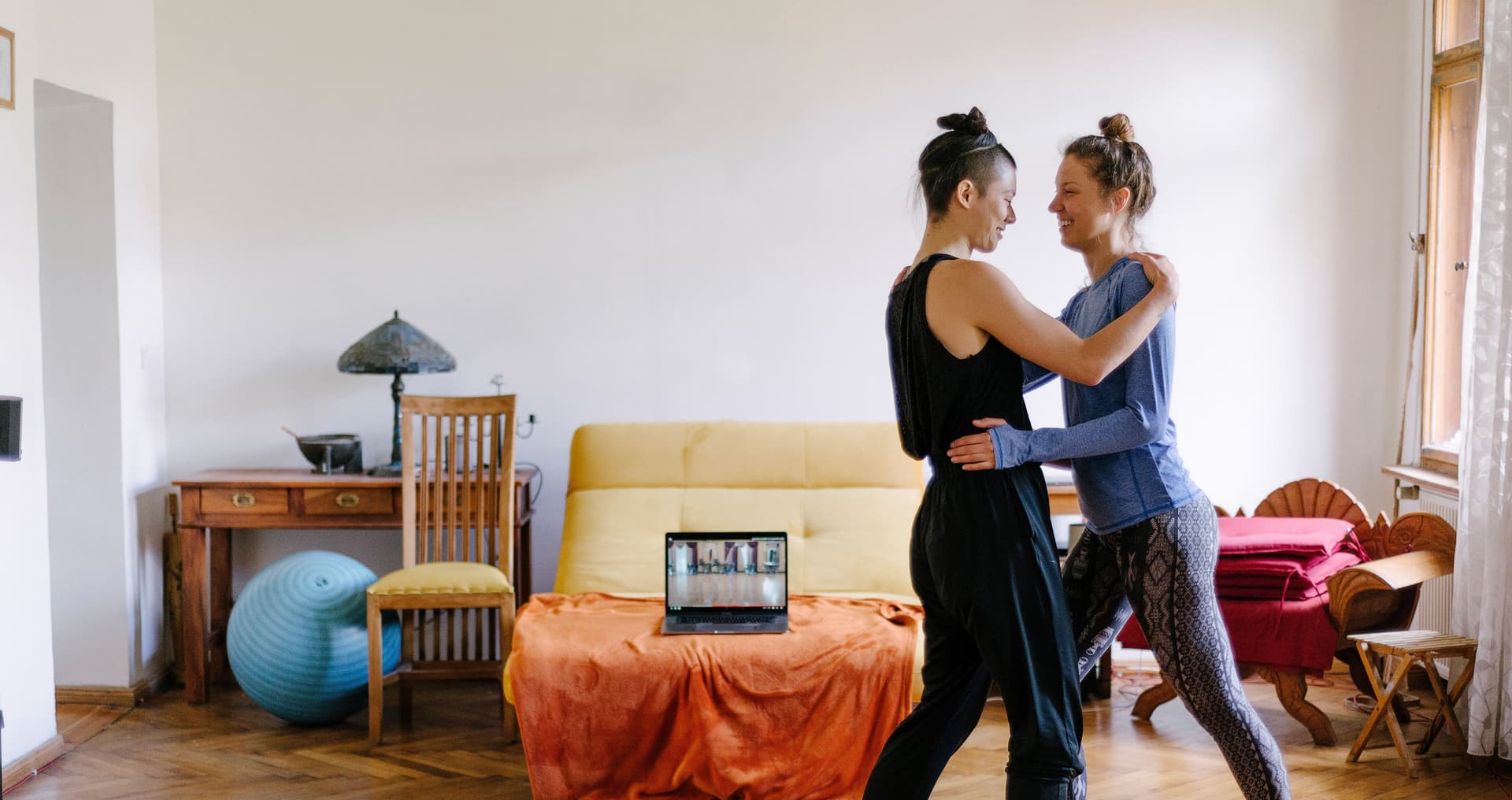 Zwei Frauen tanzen zusammen Tango in ihrer Altbauwohnung. Im Hintergrund steht ein Laptop, auf dem ein Online-Tanzkurs zu sehen ist.