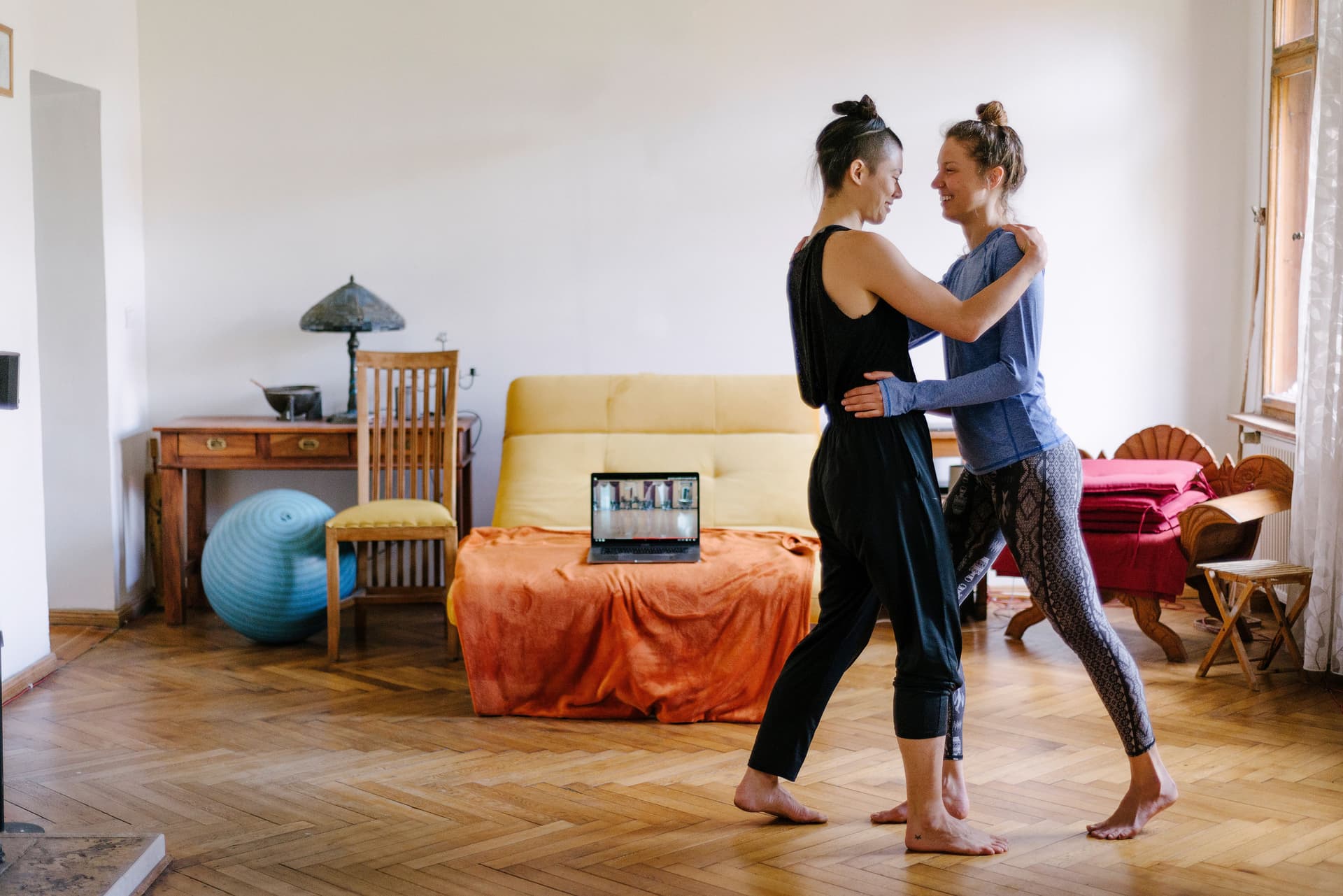 Zwei Frauen tanzen zusammen Tango in ihrer Altbauwohnung. Im Hintergrund steht ein Laptop, auf dem ein Online-Tanzkurs zu sehen ist.