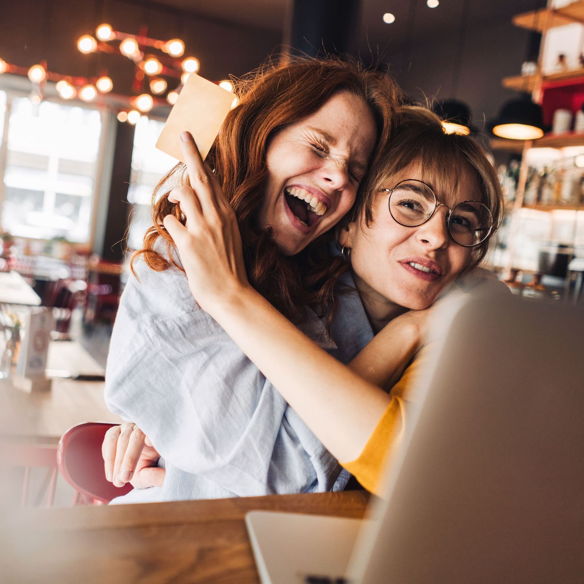 Zwei Freundinnen umarmen sich euphorisch. Sie sitzen in einem Cafe vor einem Laptop. Eine der jungen Frauen hält eine Bankkarte hoch.
