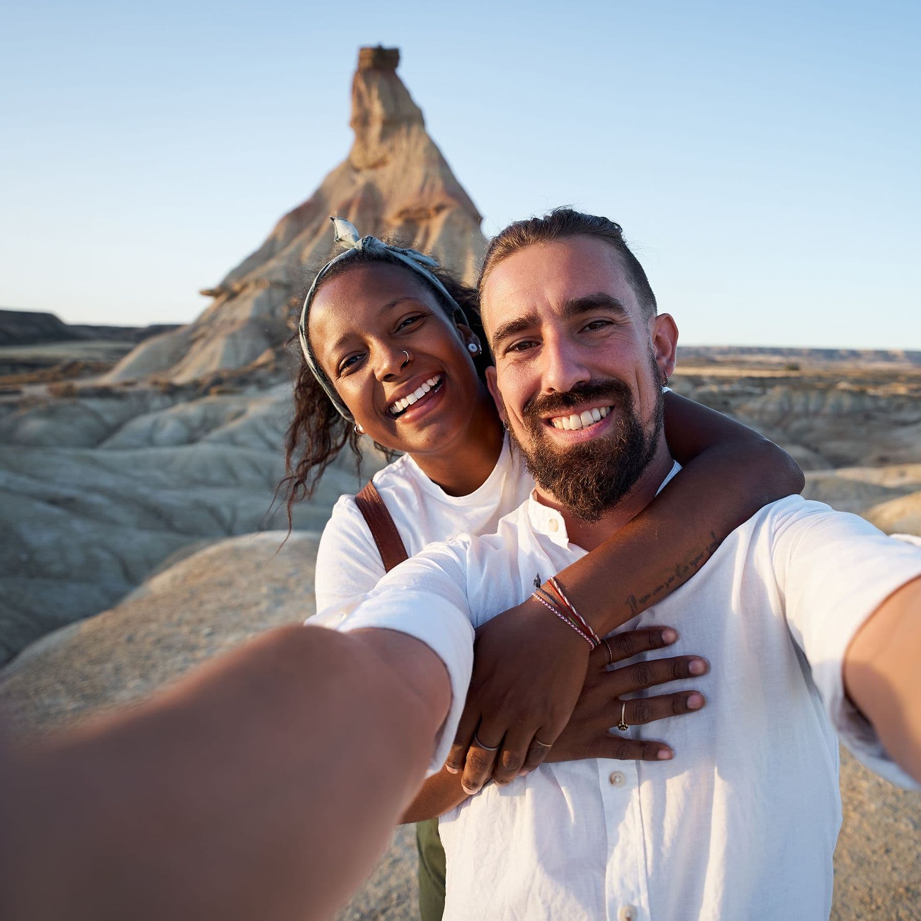 Fröhliches Selfie eines Paares in einer steinigen Wüstenlandschaft mit einem Felsberg im Hintergrund.