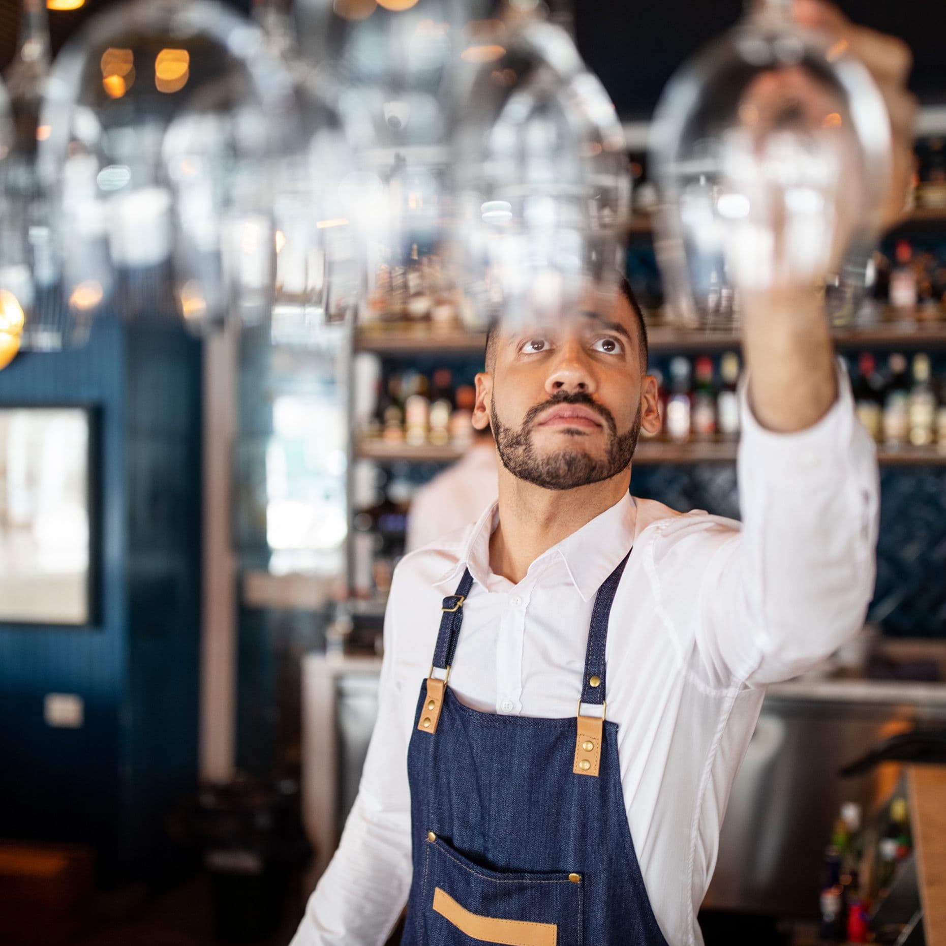 Ein Restaurantmitarbeiter mittleren Alters mit Schürze und weißem Hemd hängt ein Weinglas in einen Gläserhalter über dem Tresen. 
