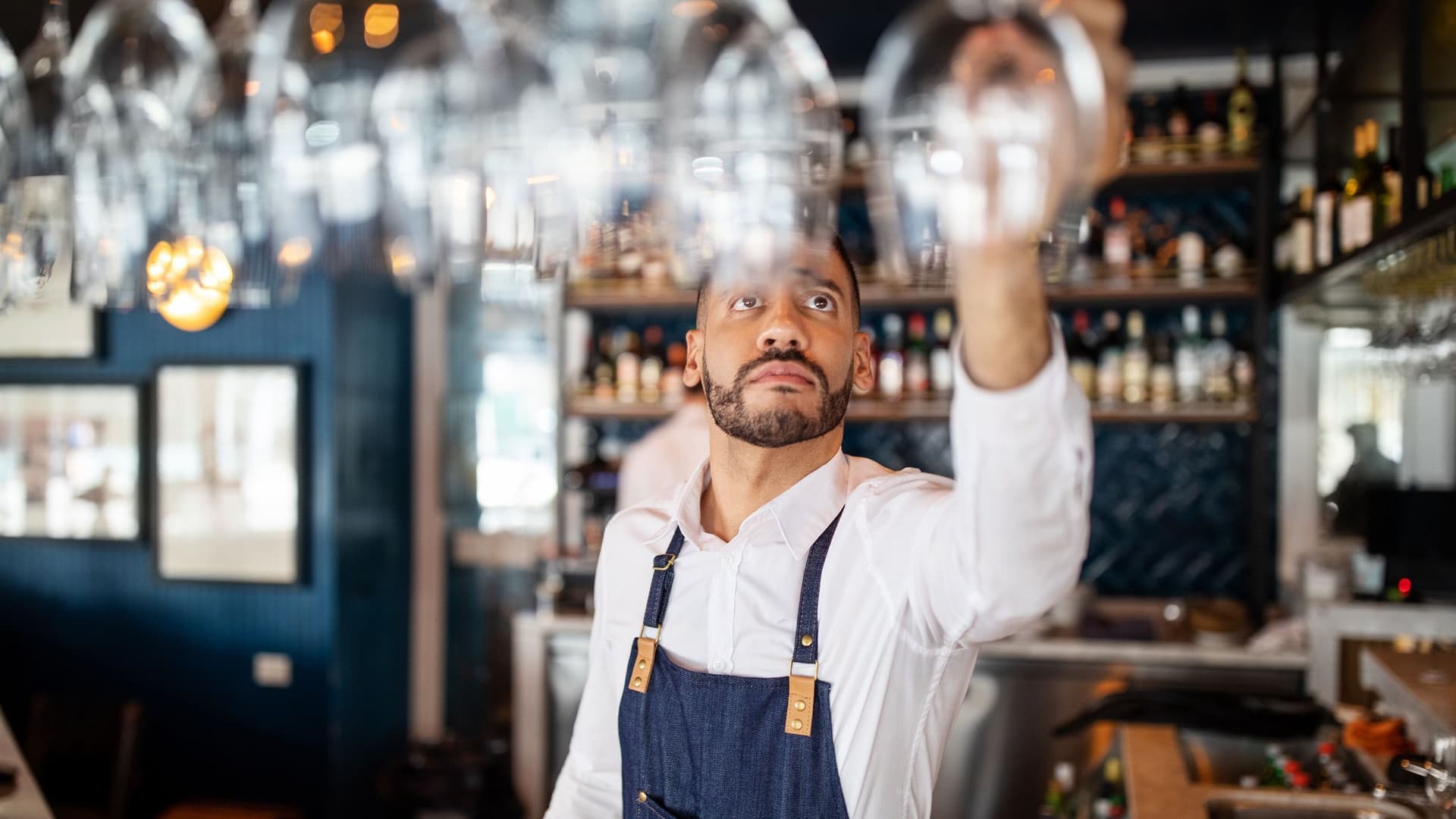 Ein Restaurantmitarbeiter mittleren Alters mit Schürze und weißem Hemd hängt ein Weinglas in einen Gläserhalter über dem Tresen. 