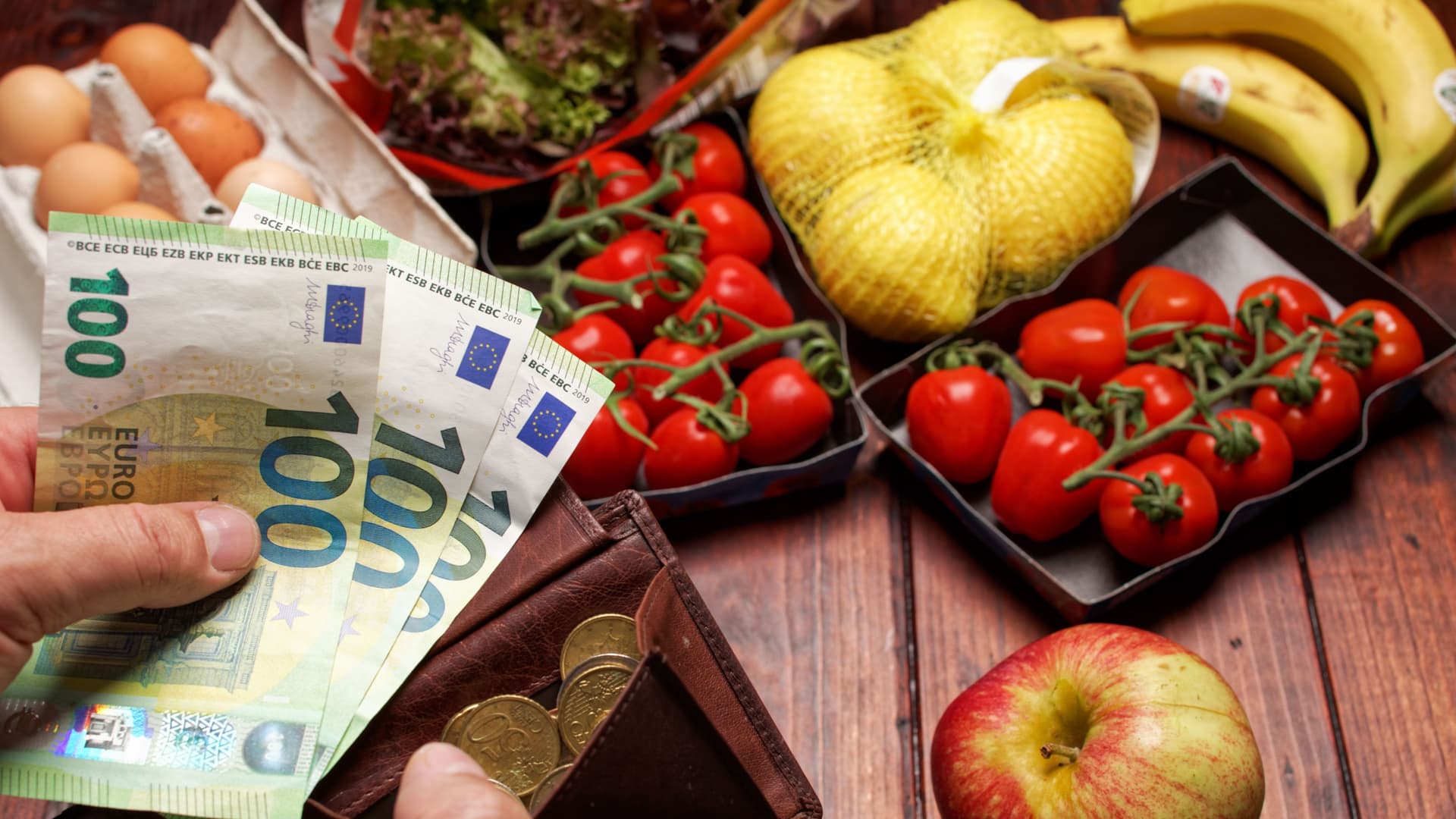 EIne Hand hält dre einhundert Euro-Scheine die andere Hand hält einen Geldbeutel im Hintergund liegt ein Einkauf mit Obst Gemüse und Eiern.