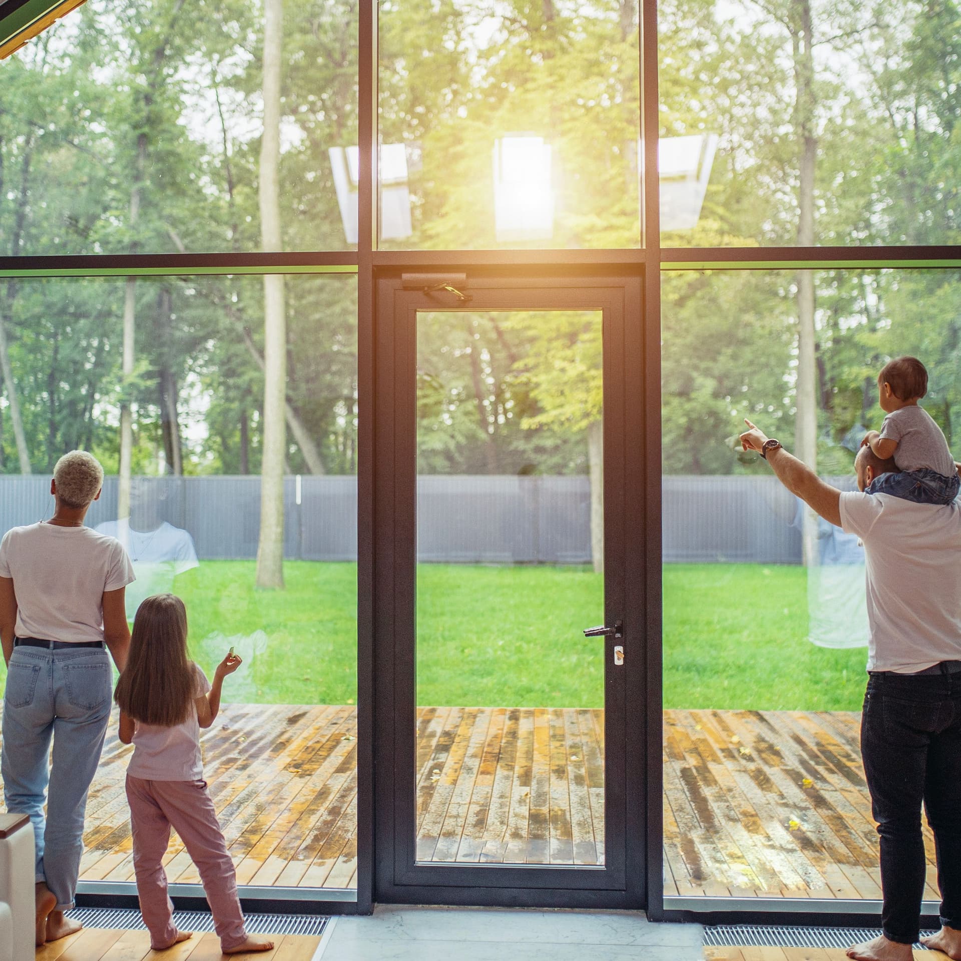 Eltern mit Kindern stehen vor einem großen Panoramafenster mit Tür und gucken in den Garten. Das Baby sitzt auf den Schultern des Vaters und die Tochter steht neben der Mutter.