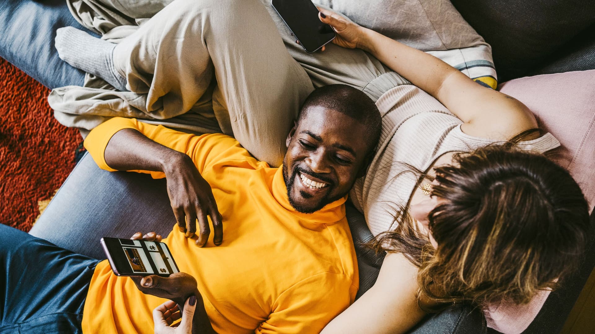 Lachender Mann mit Smartphone in der Hand, der mit seiner Freundin auf dem Sofa liegt und ihr etwas auf dem Bildschirm zeigt.