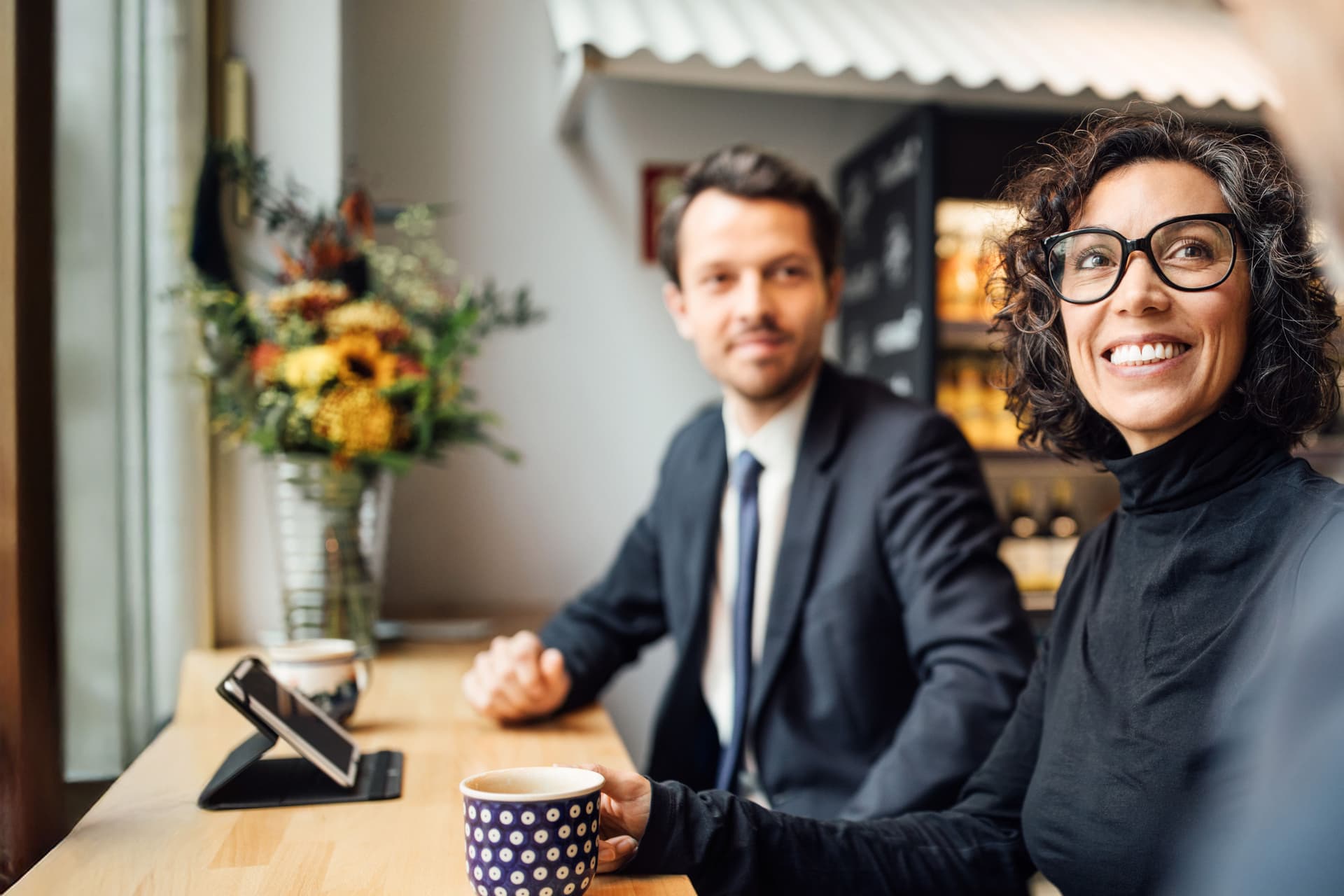 Ein Mann und eine Frau in Business-Kleidung sitzen in einem Cafe zusammen an einem langen Tisch. Sie lächeln und gucken einen anderen Mann an, den man nur zum Teil sieht.