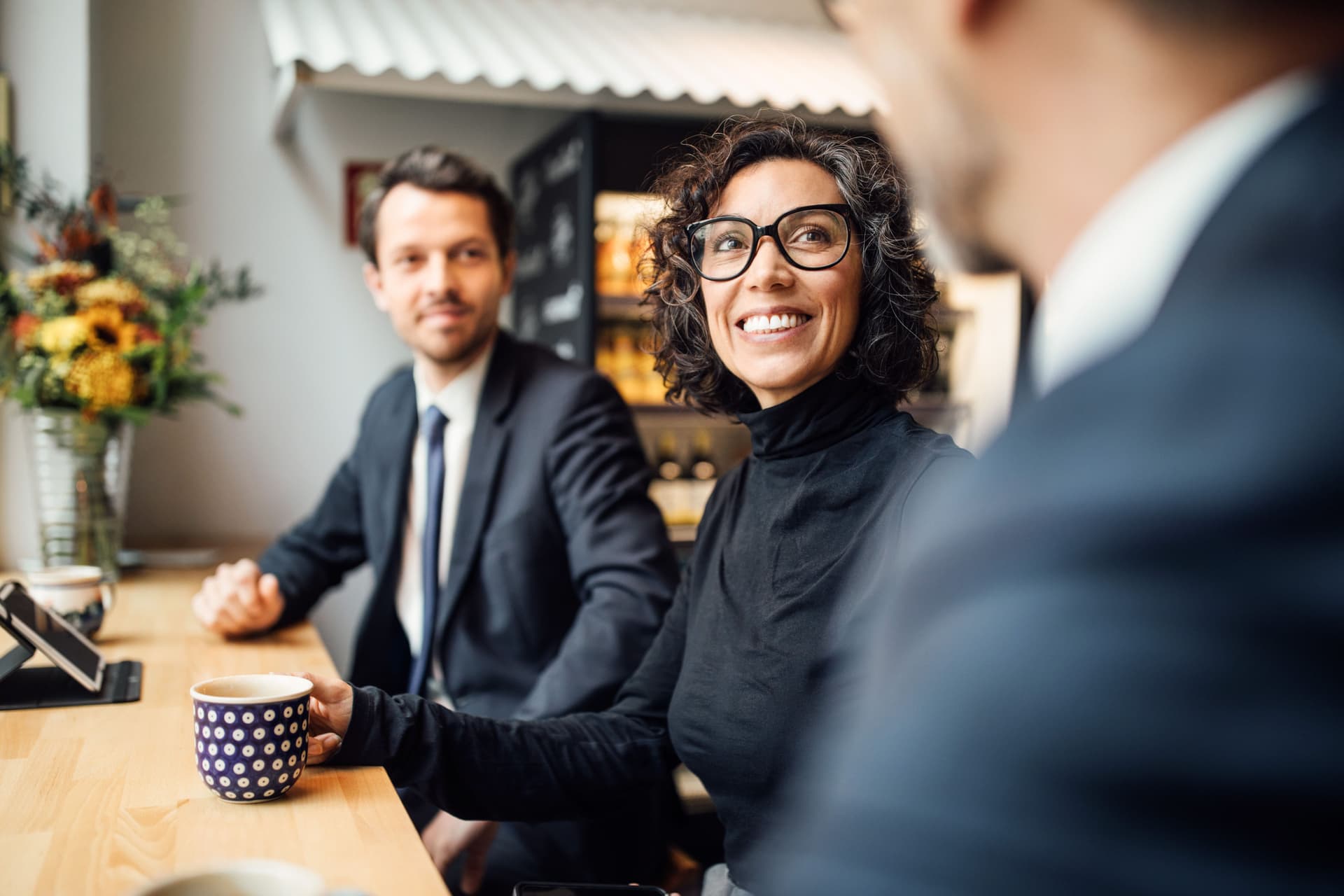 Ein Mann und eine Frau in Business-Kleidung sitzen in einem Cafe zusammen an einem langen Tisch. Sie lächeln und gucken einen anderen Mann an, den man nur zum Teil sieht.