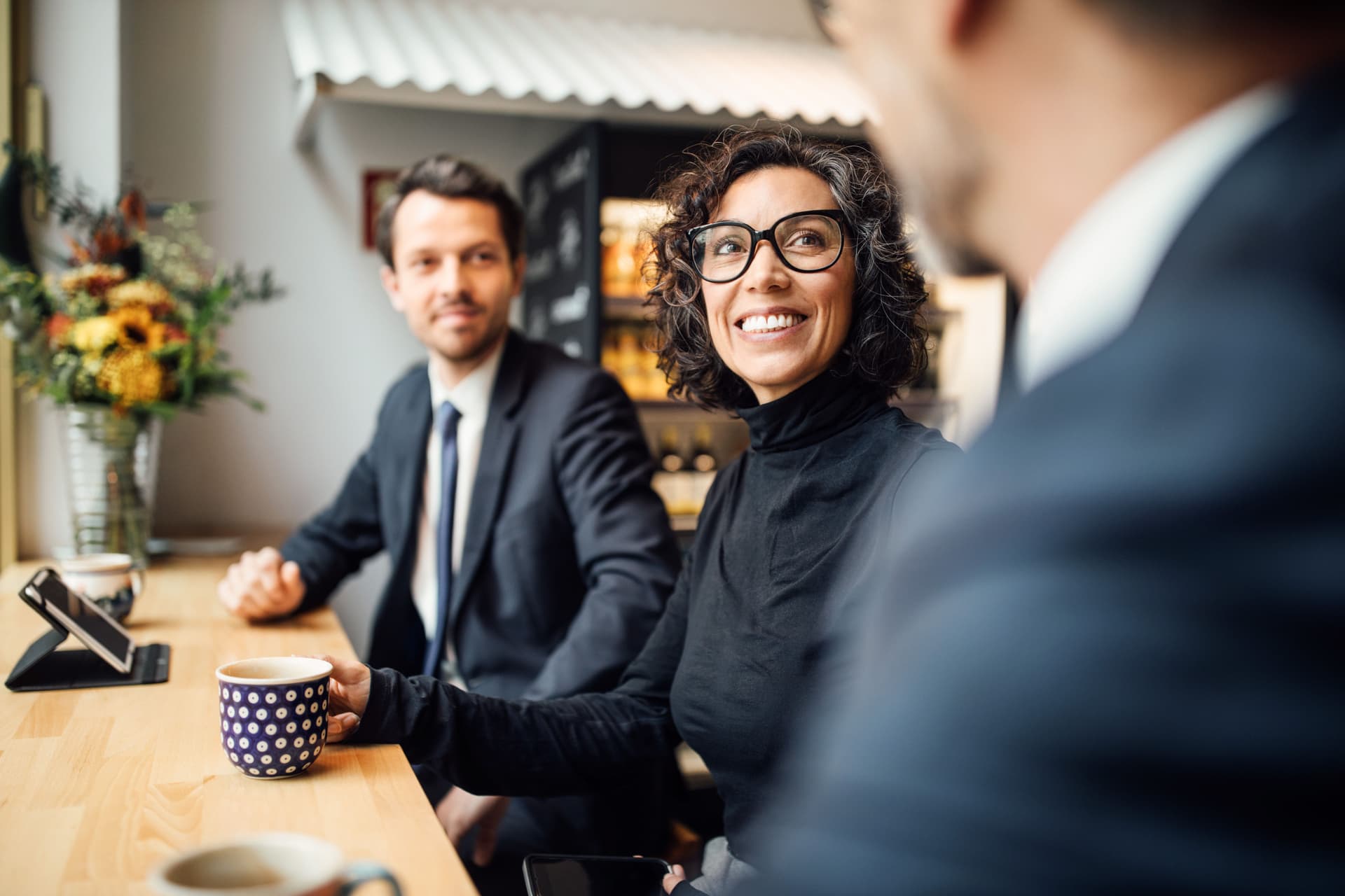 Ein Mann und eine Frau in Business-Kleidung sitzen in einem Cafe zusammen an einem langen Tisch. Sie lächeln und gucken einen anderen Mann an, den man nur zum Teil sieht.