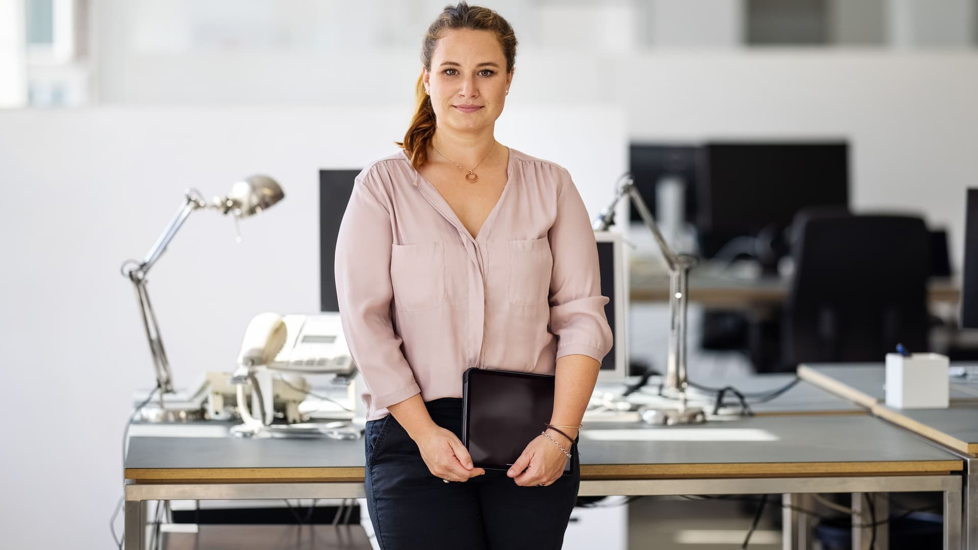 Eine junge Frau hält ein Tablet in den Händen und lehnt sich dabei an einen Schreibtisch in einem Büro. Sie blickt in die Kamera.