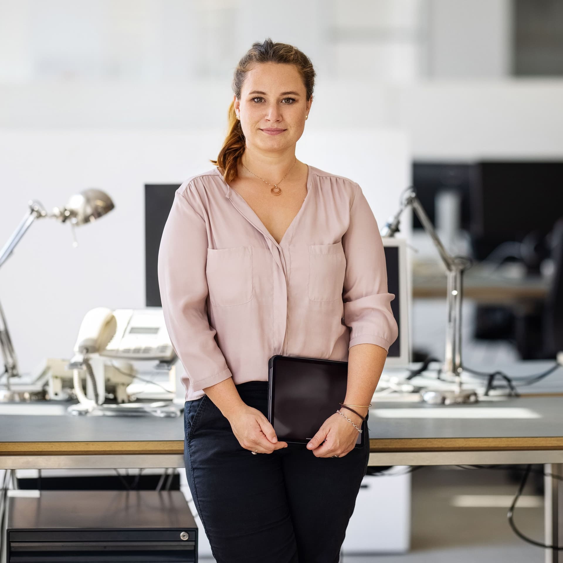 Eine junge Frau hält ein Tablet in den Händen und lehnt sich dabei an einen Schreibtisch in einem Büro. Sie blickt in die Kamera.