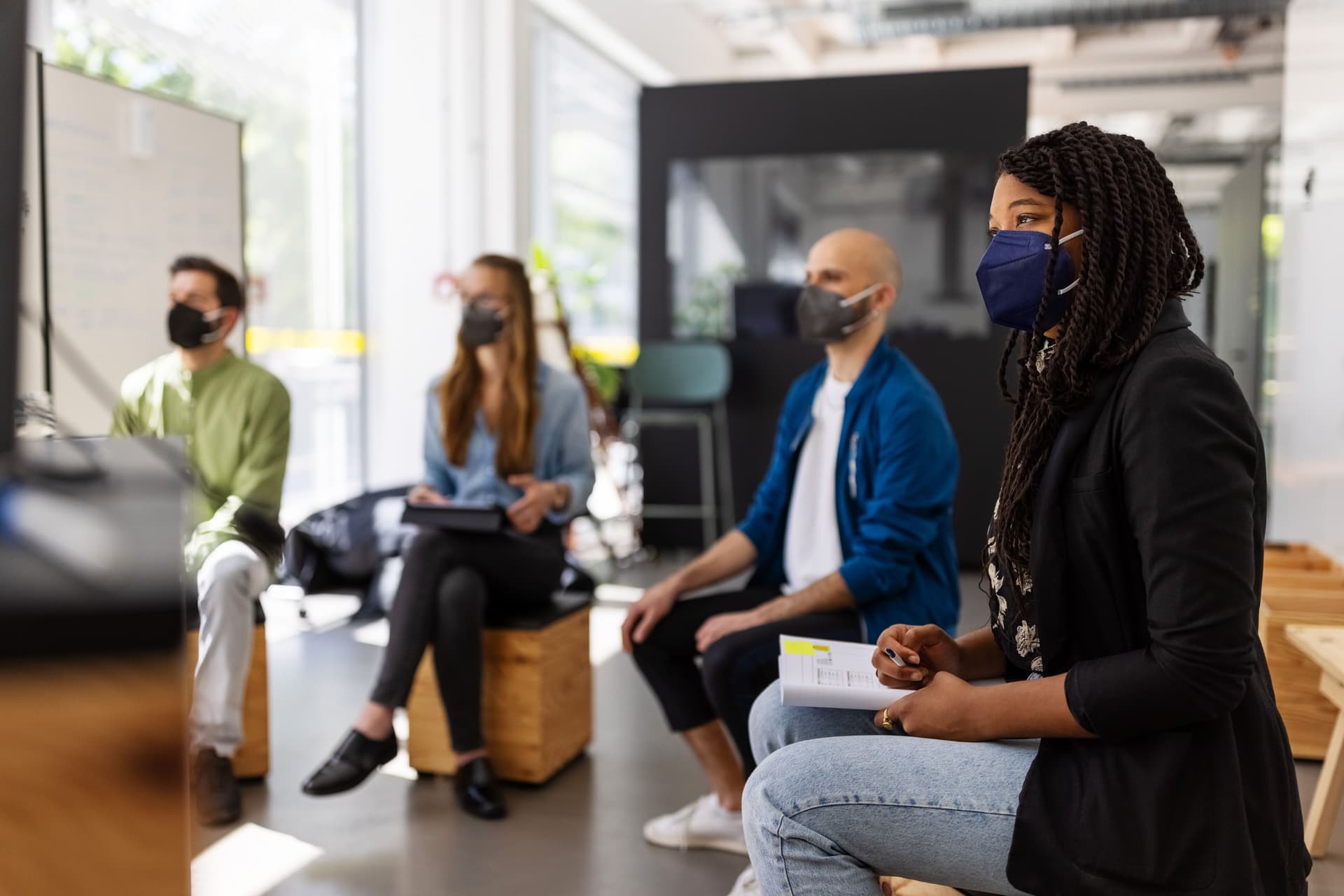 Frauen und Männer sitzen mit Gesichtsmasken auf Holzhockern in einem Büro. Sie gucken alle in eine Richtung auf einen Bildschirm.