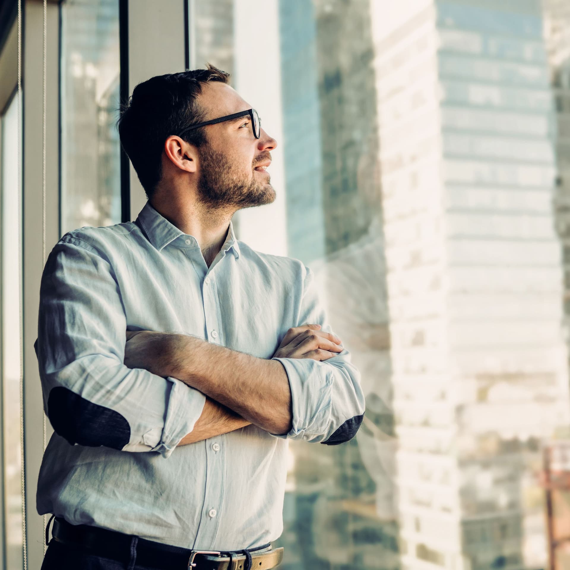 Ein junger Geschäftsmann lehnt an einem Fenster und schaut raus auf Hochhäuser.