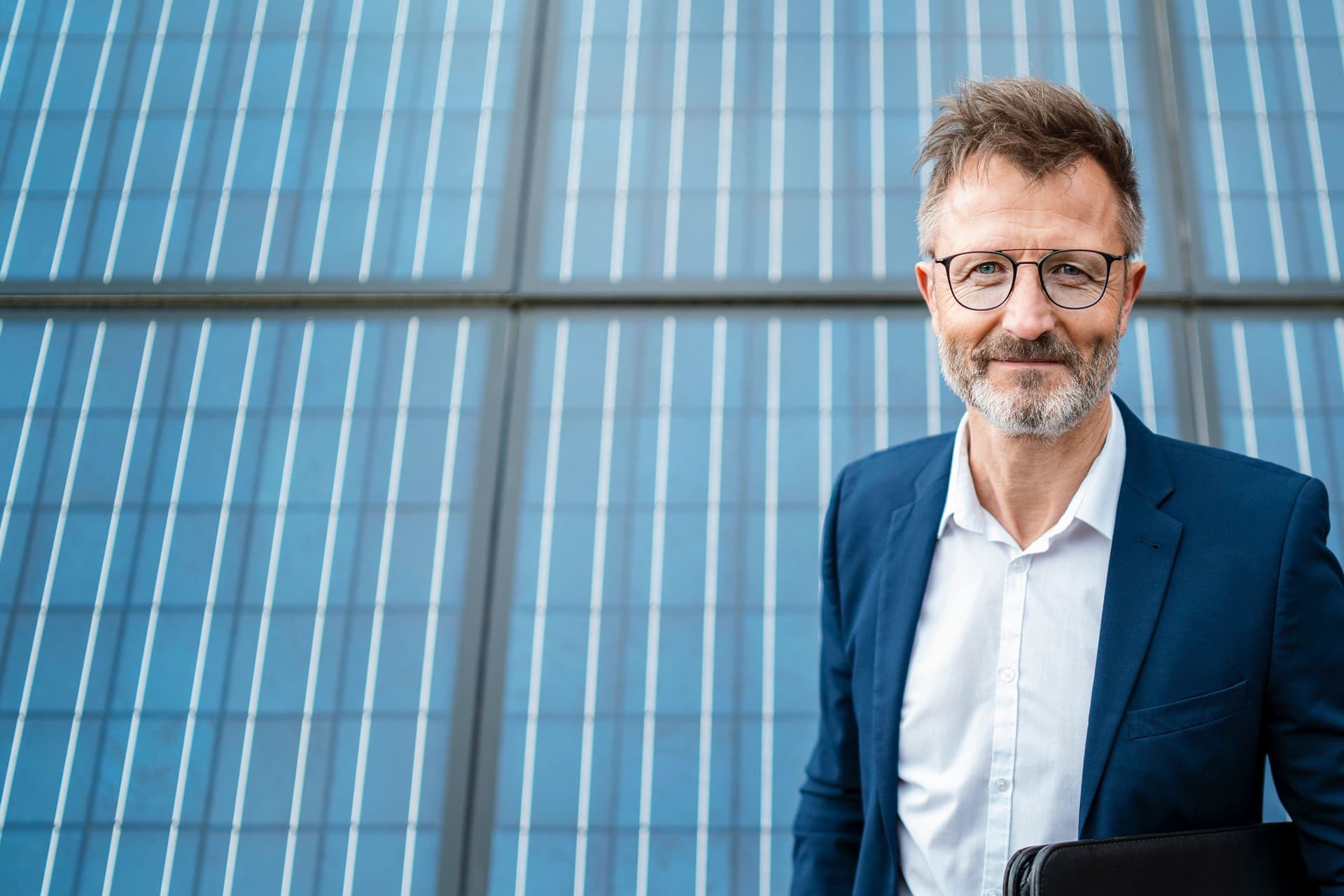 Porträt eines lächelnden Mannes, der einen Anzugjacke trägt und einen Ordner unter dem Arm hält. Er steht vor Solarzellen.