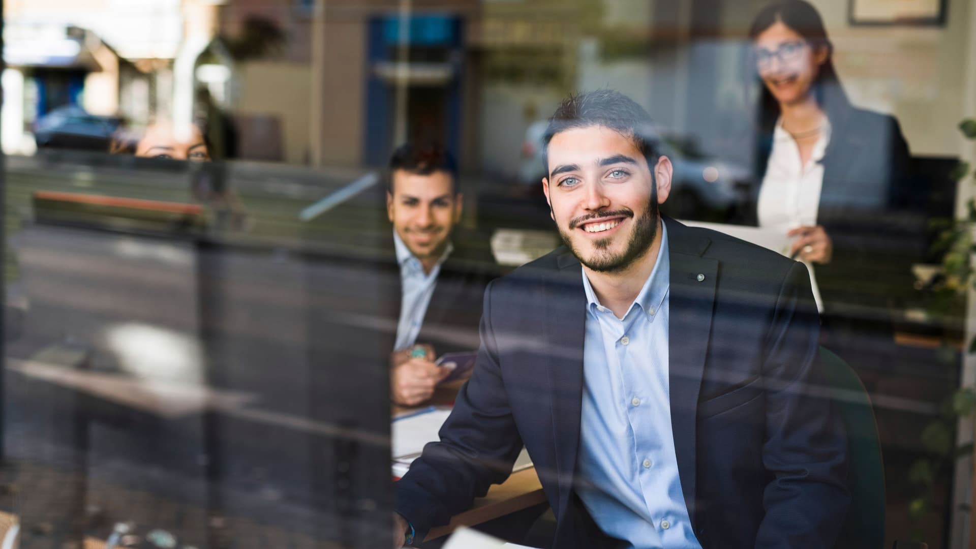 Zwei Männer und eine Frau in Businness-Kleidung in einem Büro, die durch eine Glasscheibe fotografiert wurden. Sie lächeln in die Kamera.