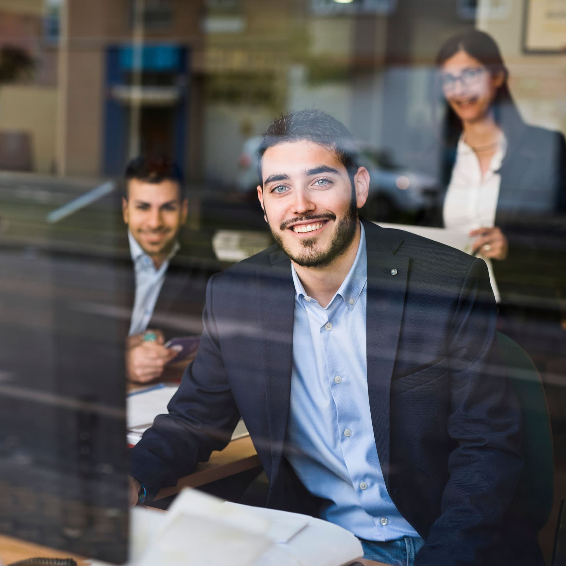 Zwei Männer und eine Frau in Businness-Kleidung in einem Büro, die durch eine Glasscheibe fotografiert wurden. Sie lächeln in die Kamera.