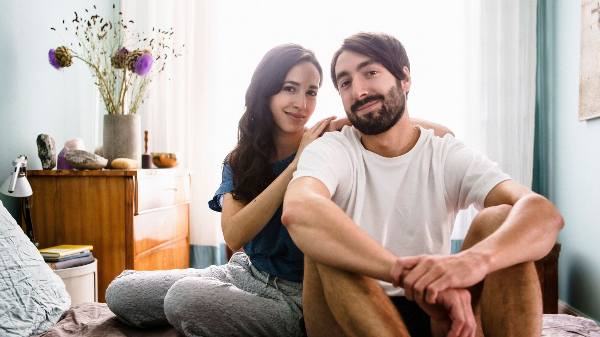 Porträt eines jungen Mannes und einer jungen Frau, die in die Kamera lächeln und zusammen auf einem Bett sitzen. Sie hat die Arme auf seine Schultern gelehnt.