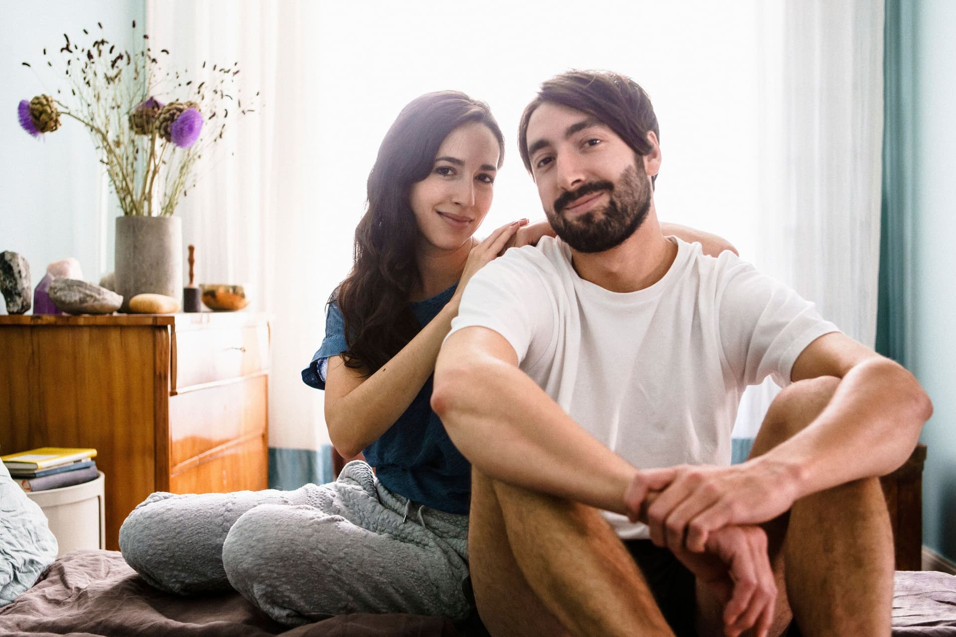 Porträt eines jungen Mannes und einer jungen Frau, die in die Kamera lächeln und zusammen auf einem Bett sitzen. Sie hat die Arme auf seine Schultern gelehnt.
