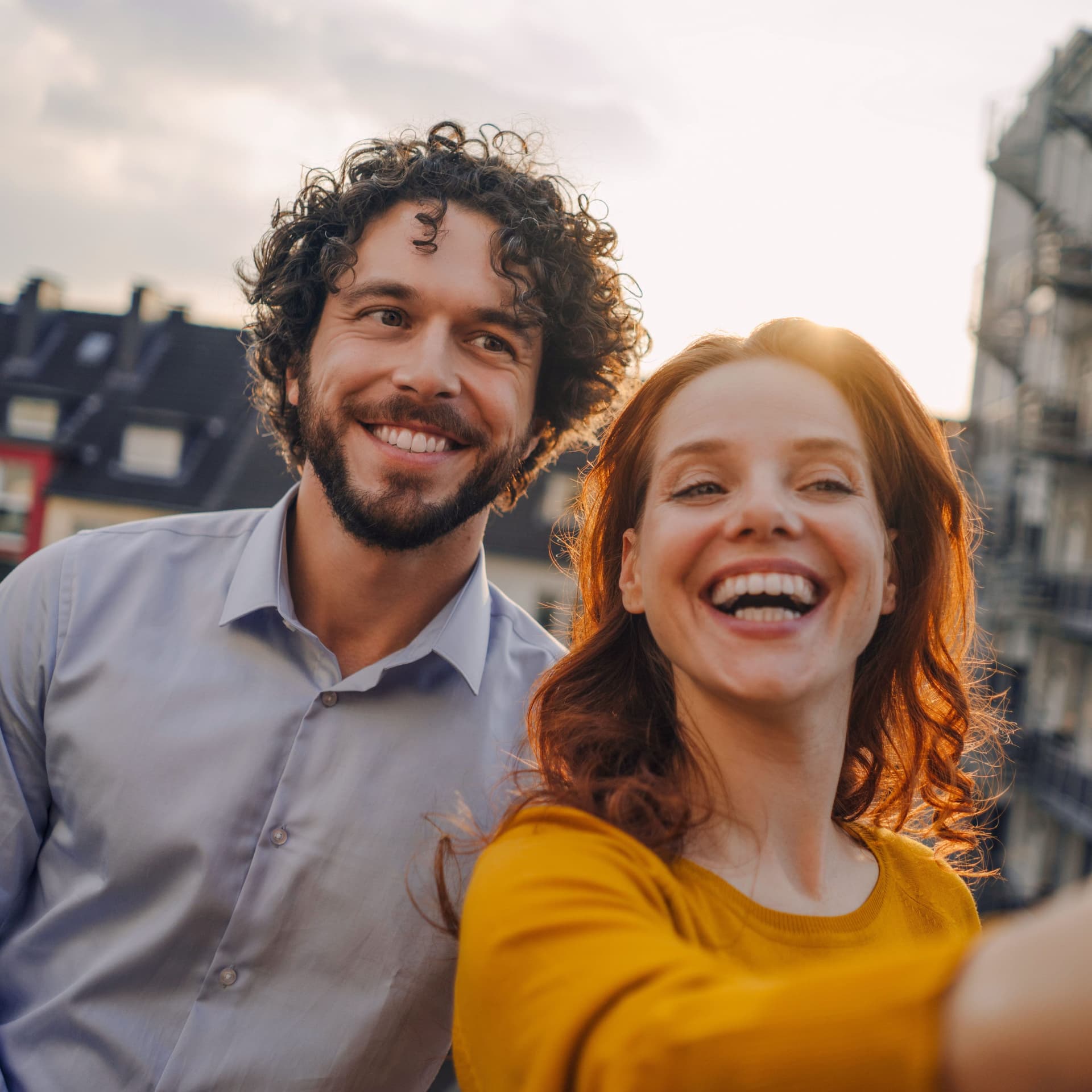 Ein Mann und eine Frau machen auf einem Balkon ein Selfie. Sie lachen ausgelassen in die Kamera. Im Hintergrund sind weitere Häuser zu sehen.